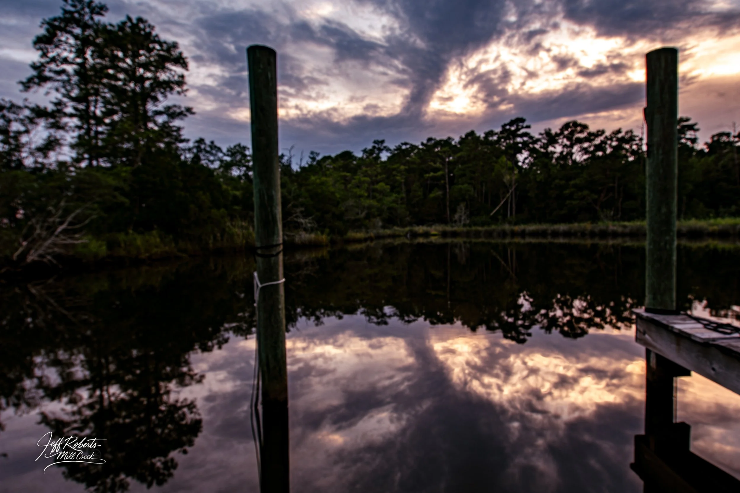 Sunset over a calm body of water reflecting the cloudy sky and trees, with wooden posts and a dock structure in the foreground.
