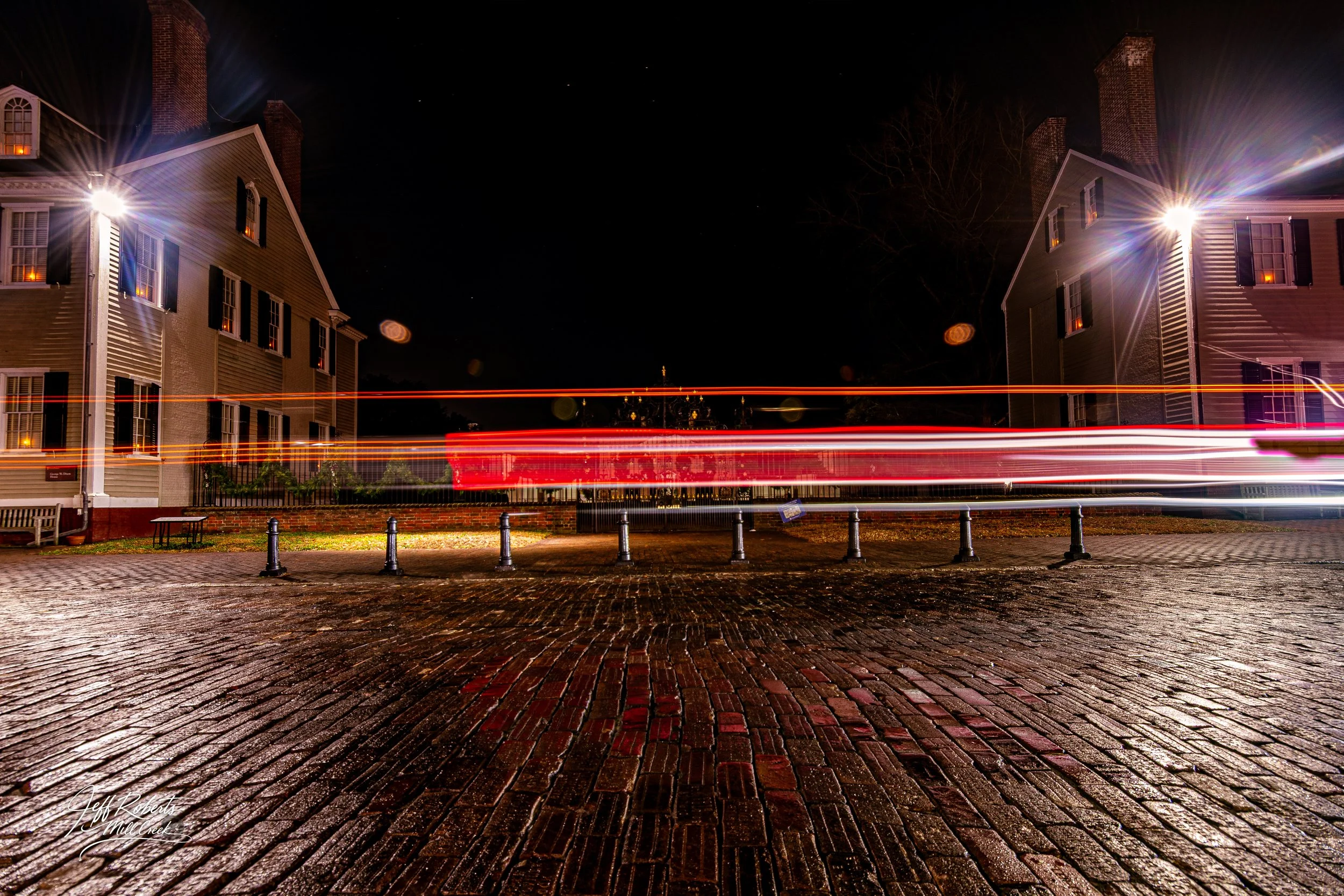 Nighttime street scene with two brick houses illuminated by bright exterior lights. Long exposure light trails from passing vehicles create horizontal streaks across the image. Wet brick street in the foreground reflects the lights, and black bollard