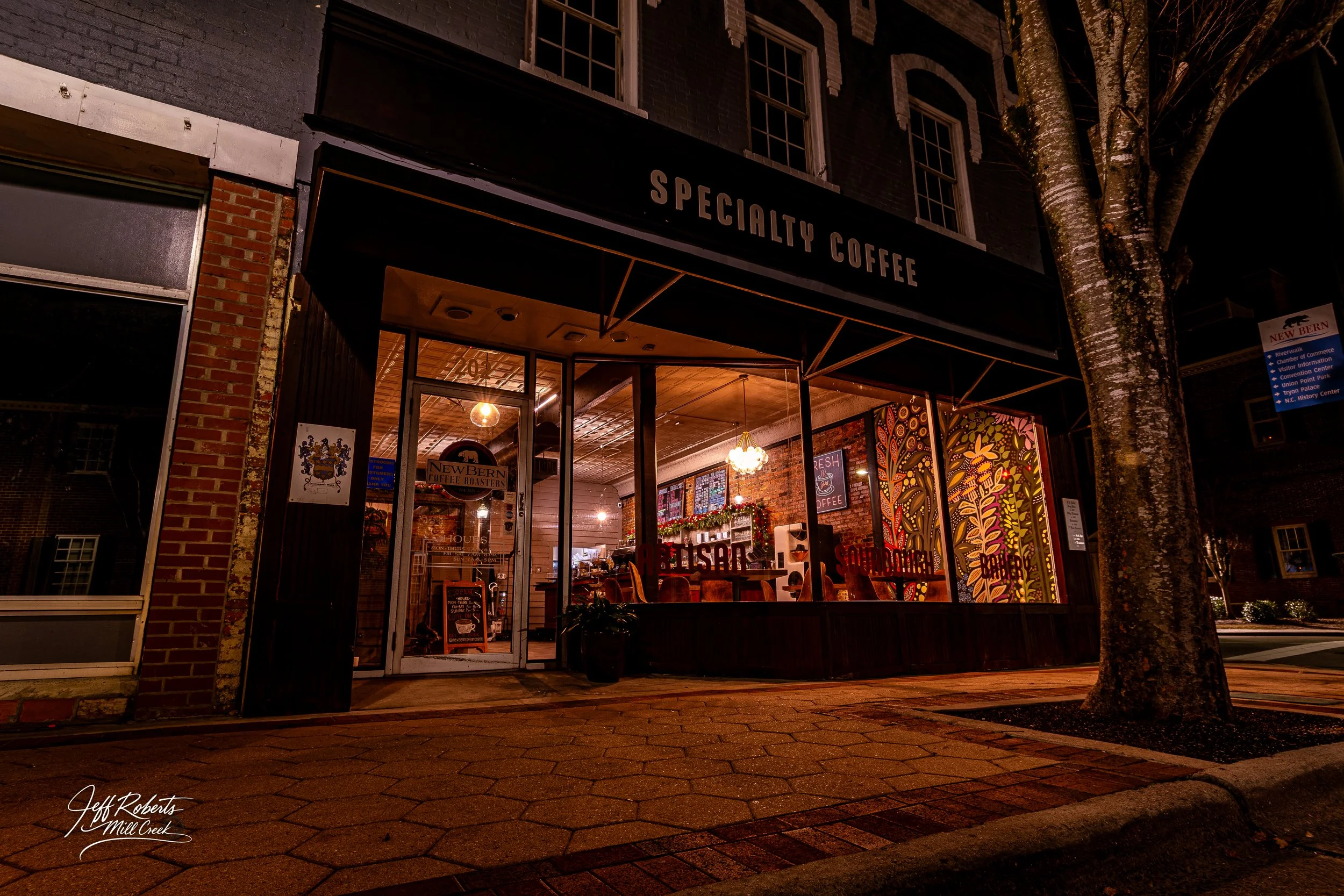 Night view of a cozy specialty coffee shop with large glass windows, decorative interior lighting, and colorful wall art inside. The shop has a black awning with 'SPECIALTY COFFEE' written on it, and a brick sidewalk outside.