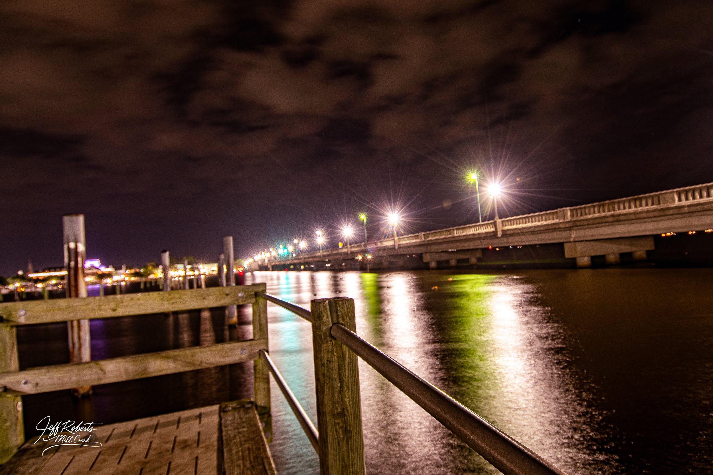 Nighttime view of a bridge over water with bright streetlights reflecting on the water, and a wooden dock with a railing in the foreground.