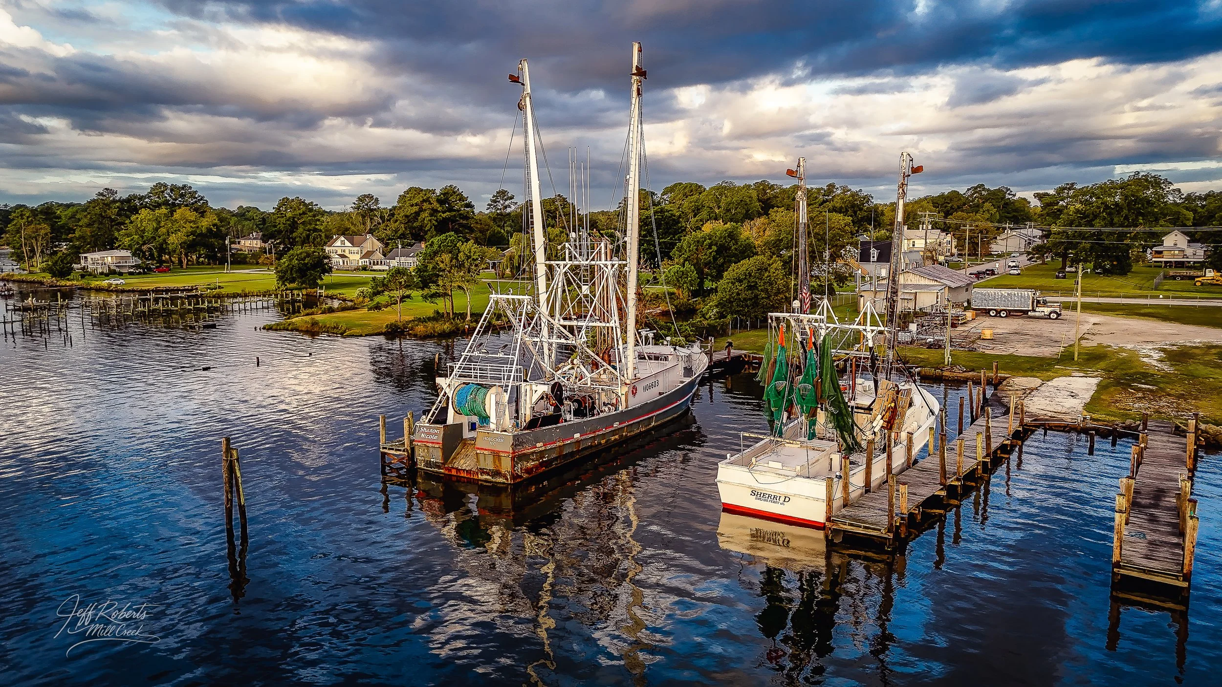 Docks with two fishing boats on a calm waterway, with a residential area and trees in the background under a cloudy sky.