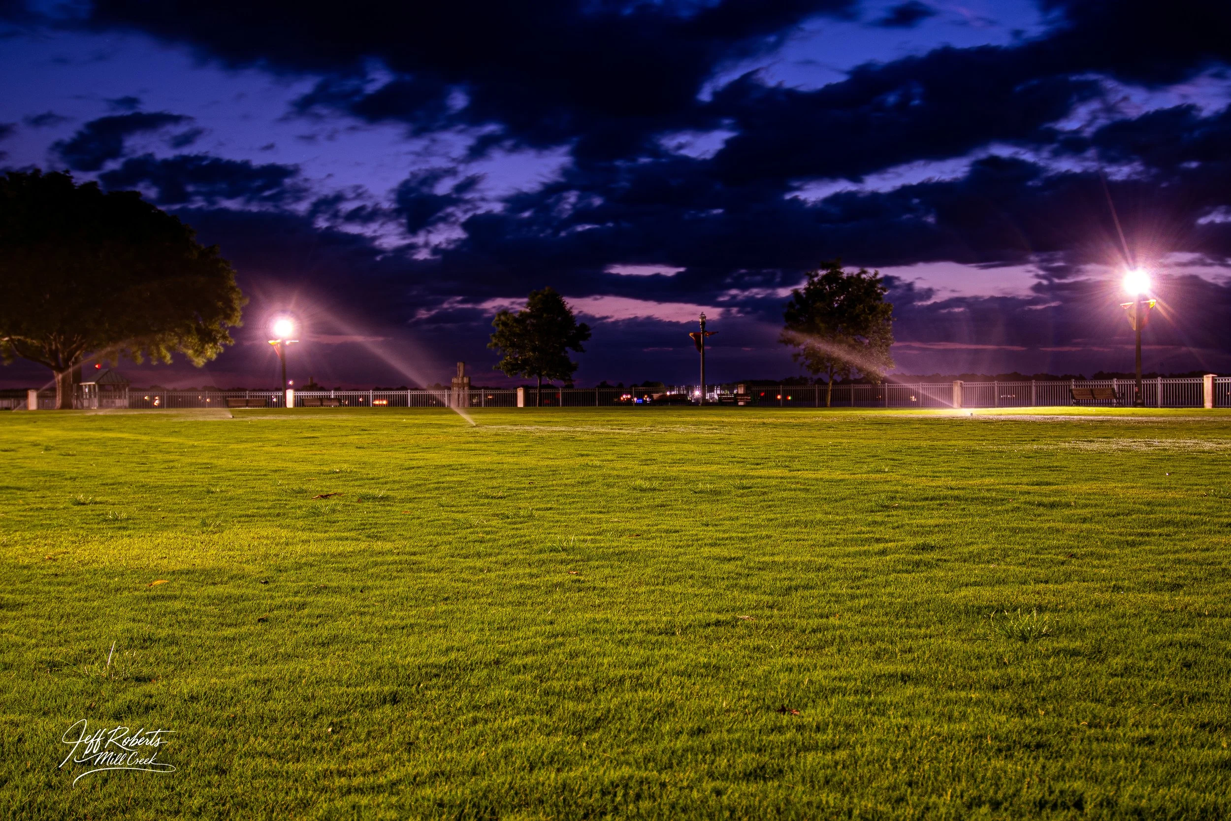 Nighttime view of a grassy park with three trees, lit by street lamps, with a cloudy sky in the background.