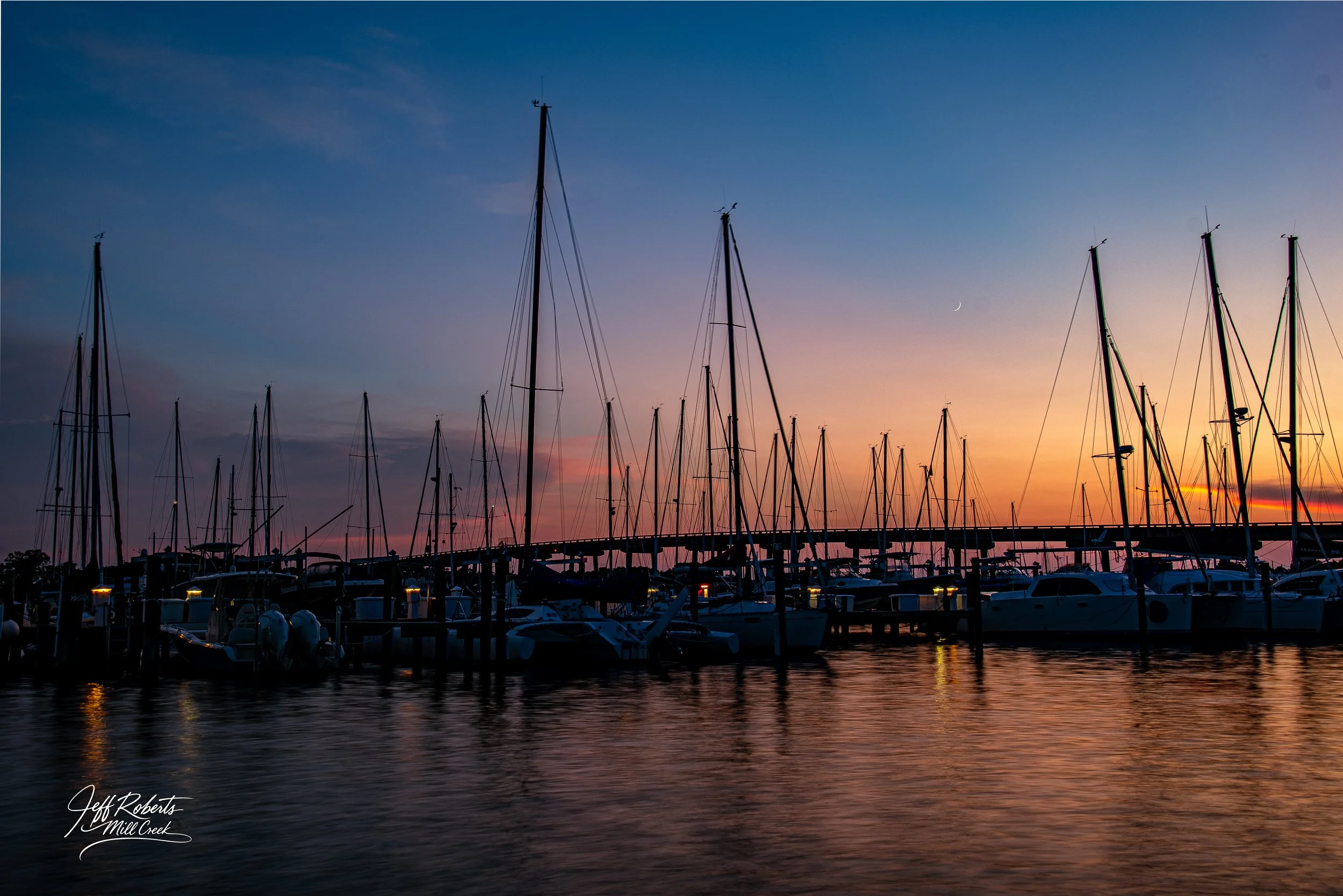 A marina at sunset with sailboats docked, colorful sky in shades of blue, pink, and orange, and a crescent moon visible in the sky.