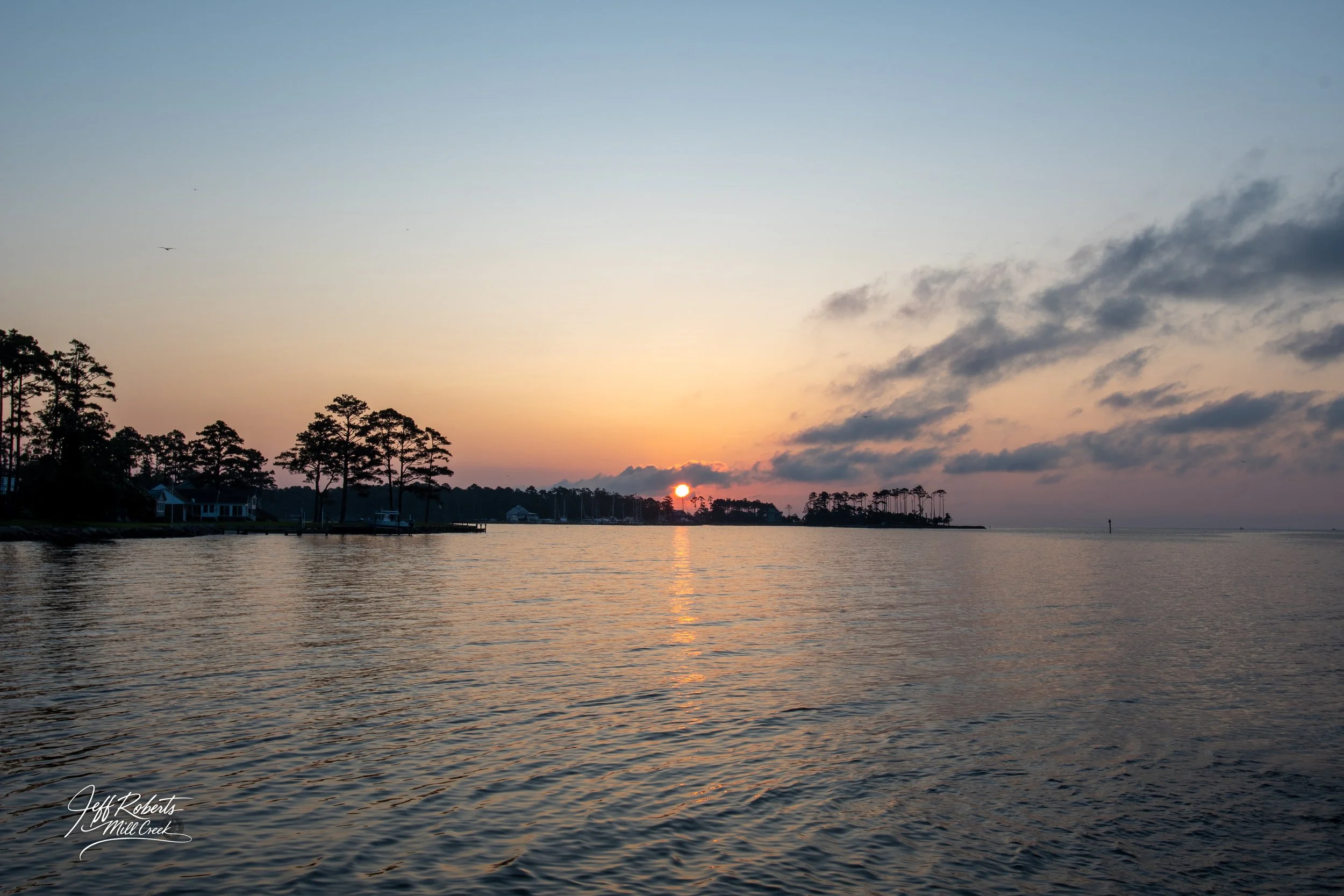Serene sunset over a calm body of water, with silhouetted trees and a few houses along the shoreline.