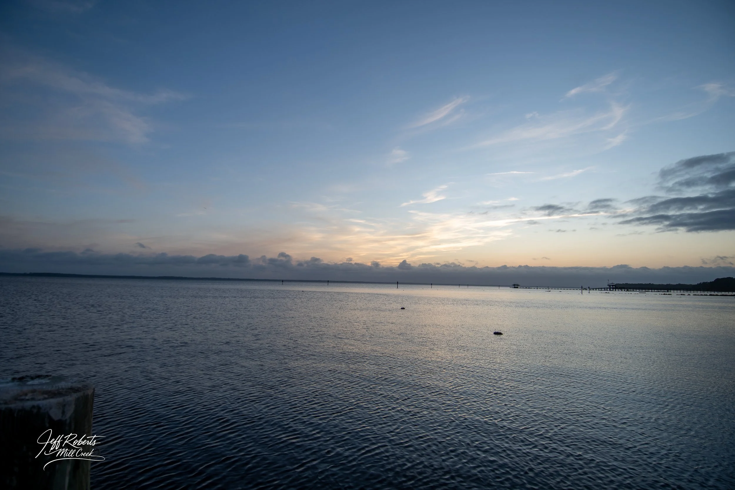 Sunset over a calm body of water with a pier in the distance and a partly cloudy sky.