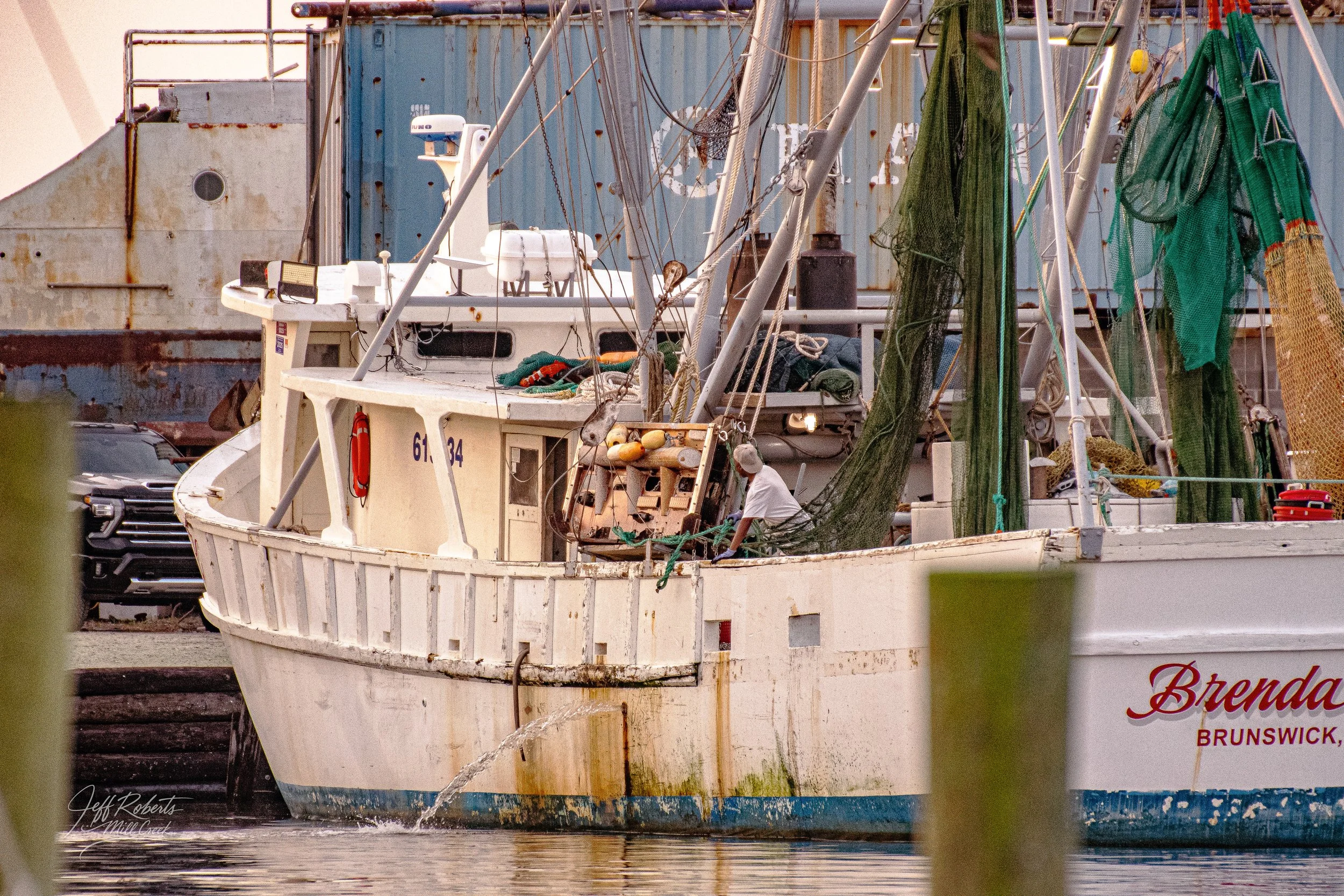 A white fishing boat named Brenda docked at a harbor, with fishing nets and equipment on deck, and an individual working near the side of the boat.