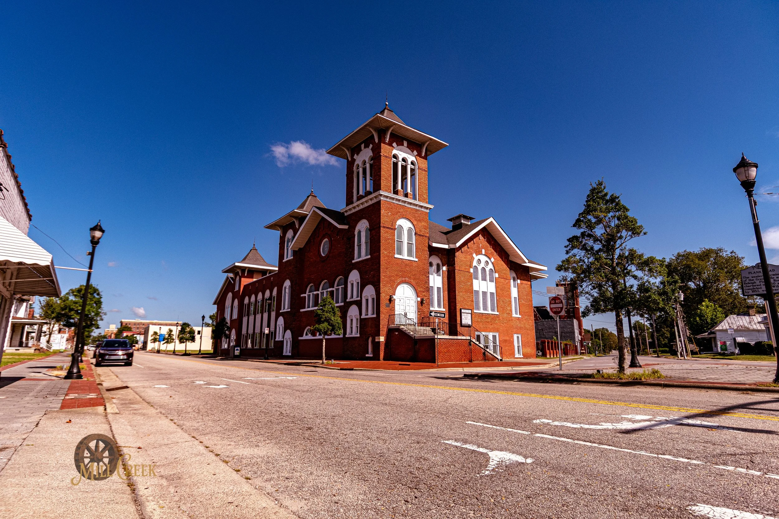 A historic red brick building with a tall tower, large arched windows, and a staircase, situated on a street corner under a clear blue sky, with trees lining the sidewalk.