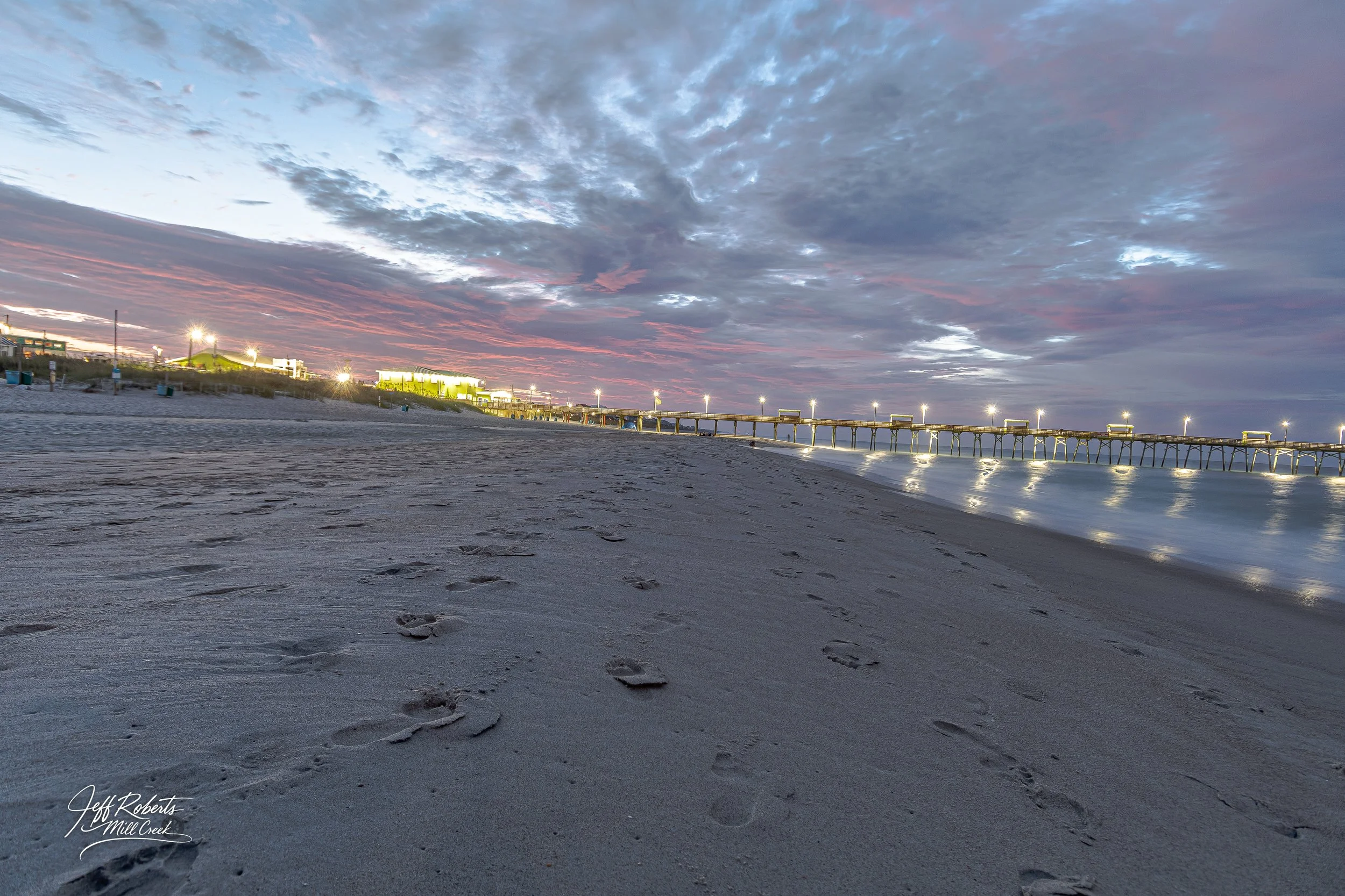 Footprints in the sand on a beach at dusk, with a pier and colorful clouds in the sky.