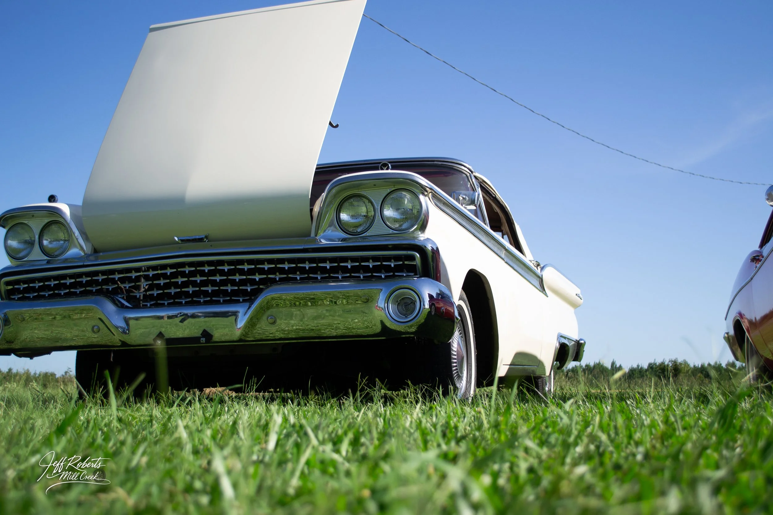A vintage white car with its hood open, parked on green grass under a clear blue sky.