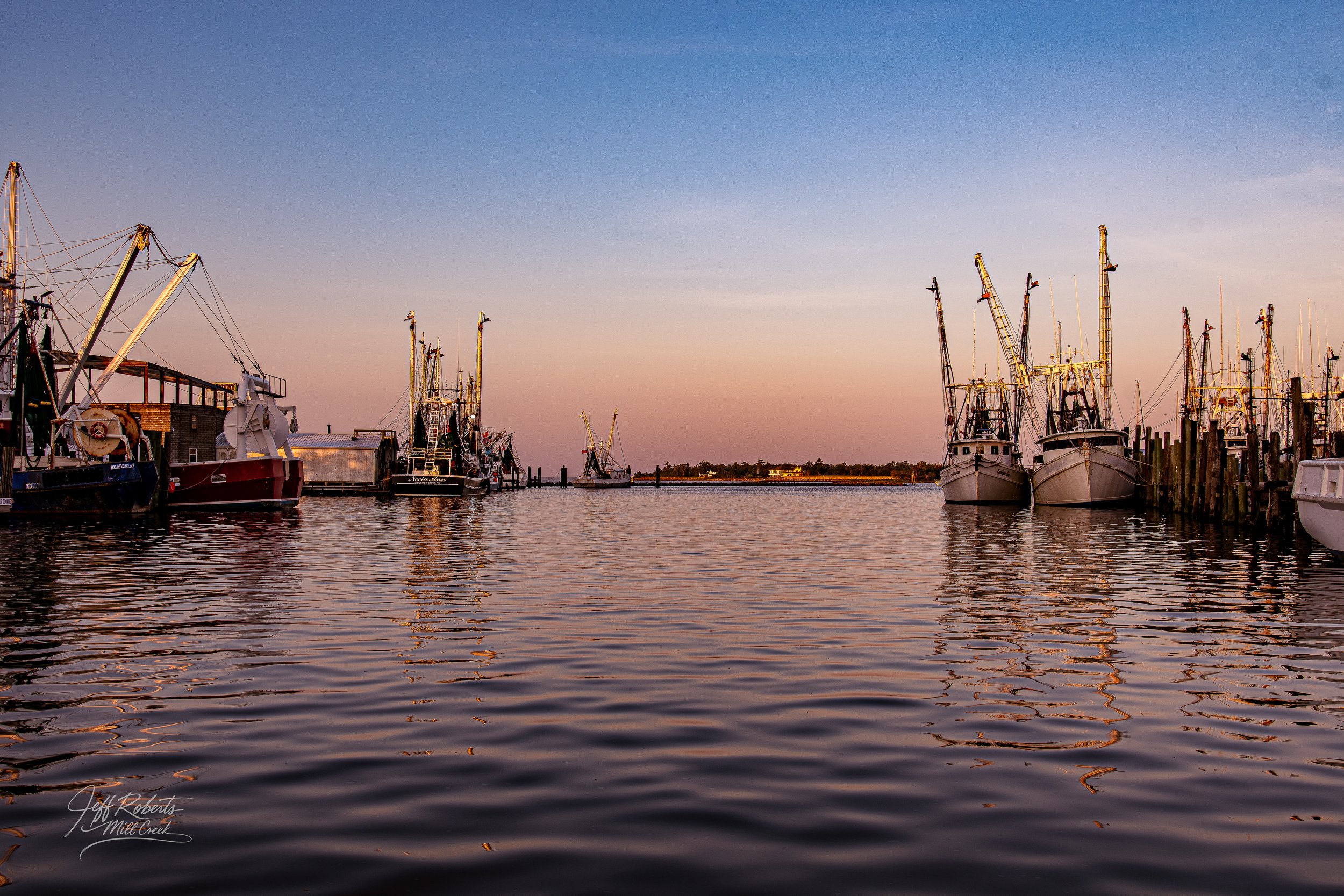 Boats docked at a marina during sunset, with calm water reflecting the boats and soft pastel sky.