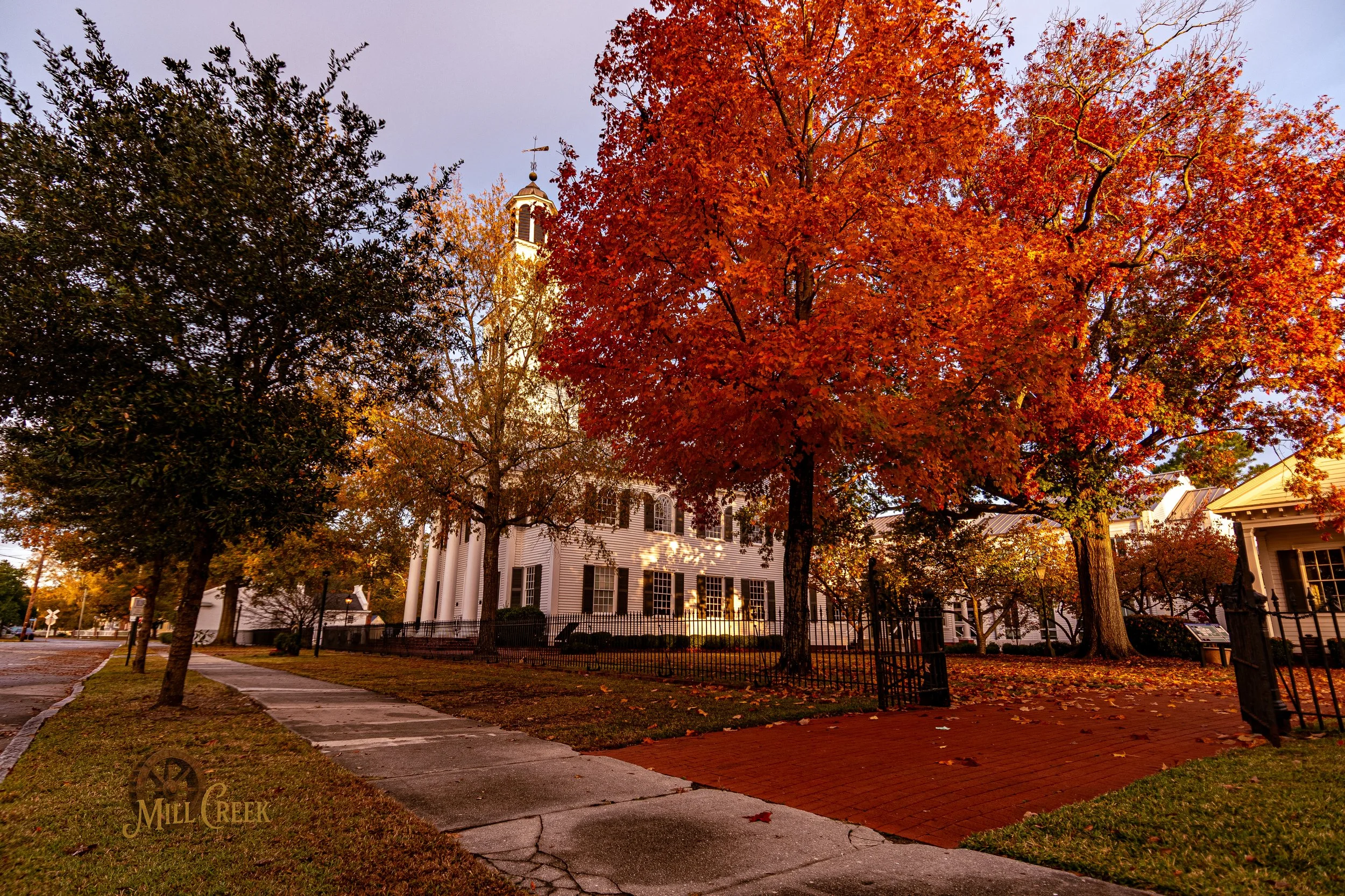 A church building with a white facade, surrounded by vibrant fall trees with orange, red, and yellow leaves, in a peaceful neighborhood setting.