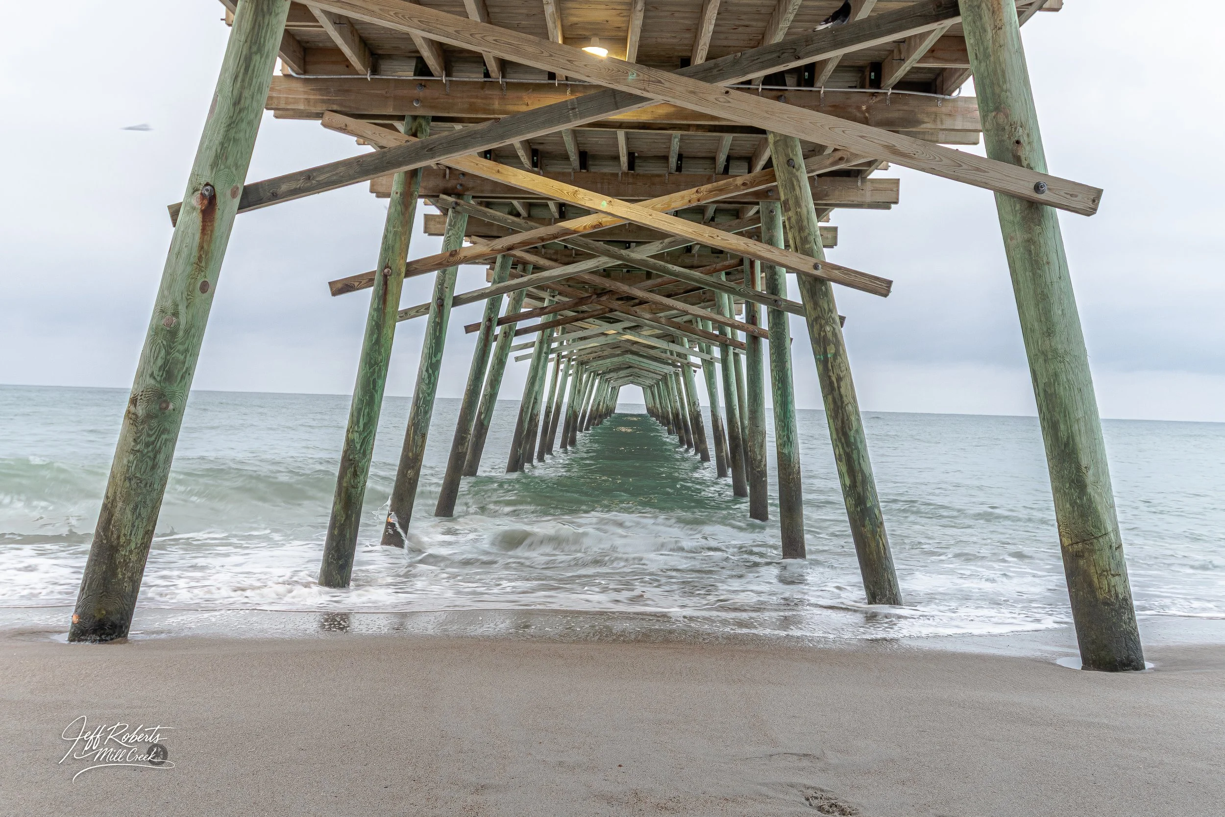 Underneath a wooden pier extending into the ocean, supported by green weathered pilings, with waves gently washing onto the sandy beach.