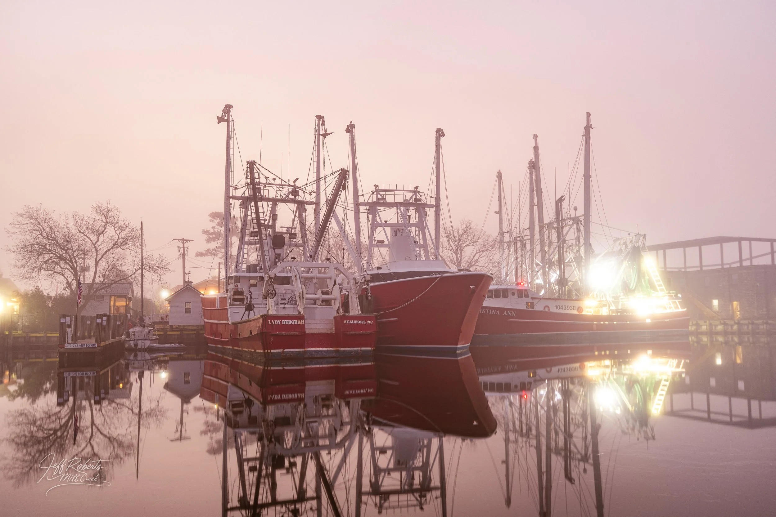 Foggy scene at a marina with several boats docked, their reflections visible in calm water, with bare trees and lamps illuminated in the background.