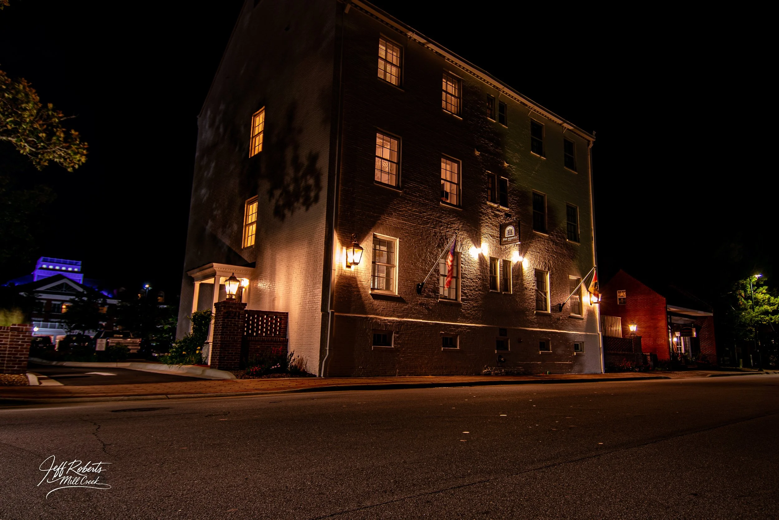 Nighttime view of a large brick building with lit windows and exterior lights, flags hanging on the front, and street in foreground.