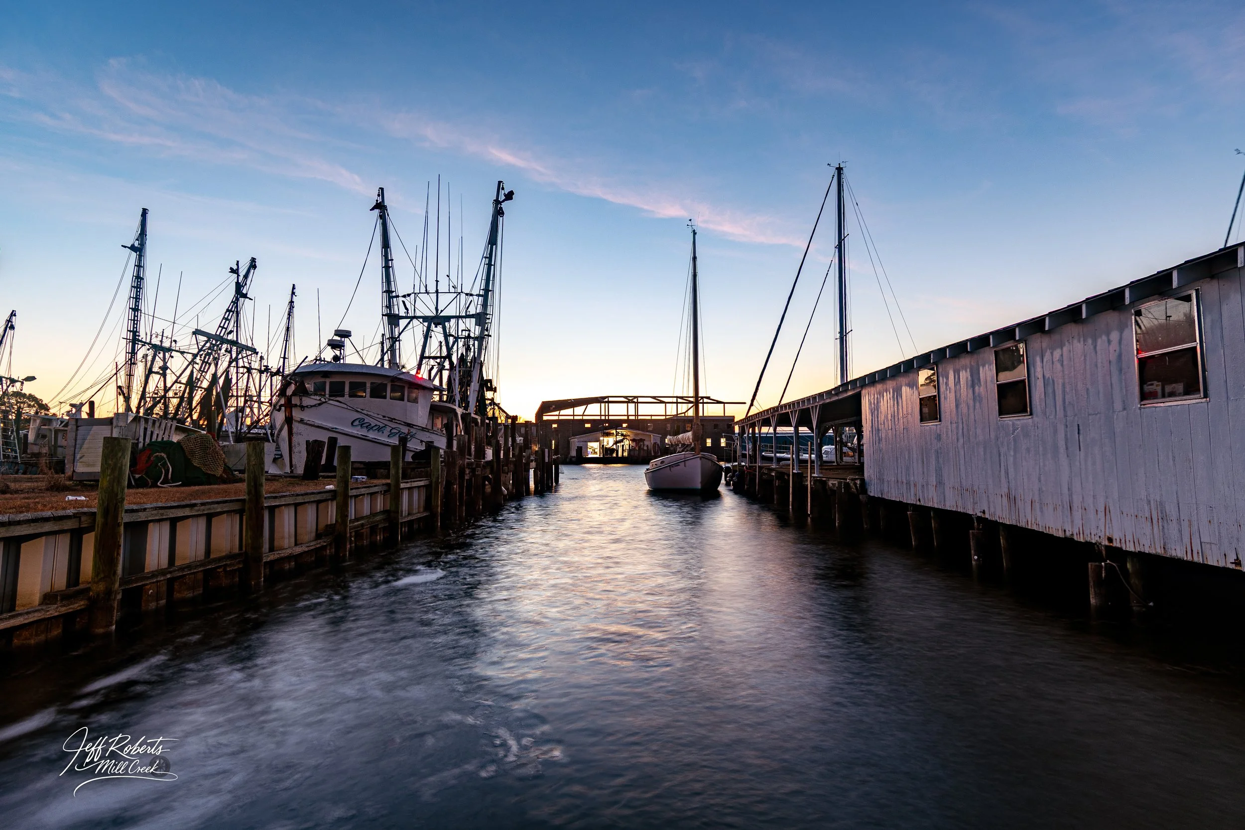 Boat dock at sunset with multiple boats moored on both sides, calm water, and a small building or shed on the right, in a marina.