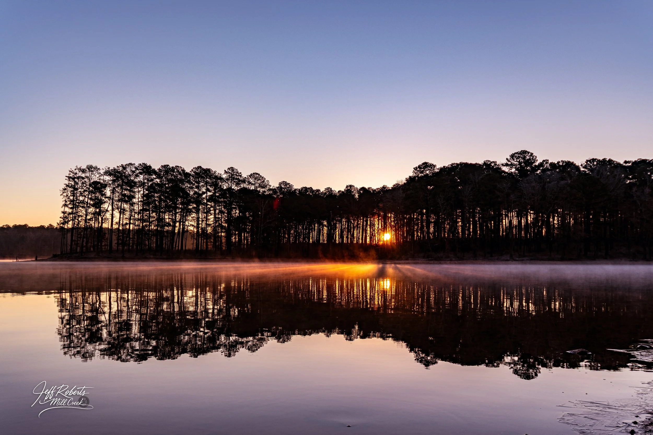 Sunrise over a lake with mist, reflecting trees and a clear sky.