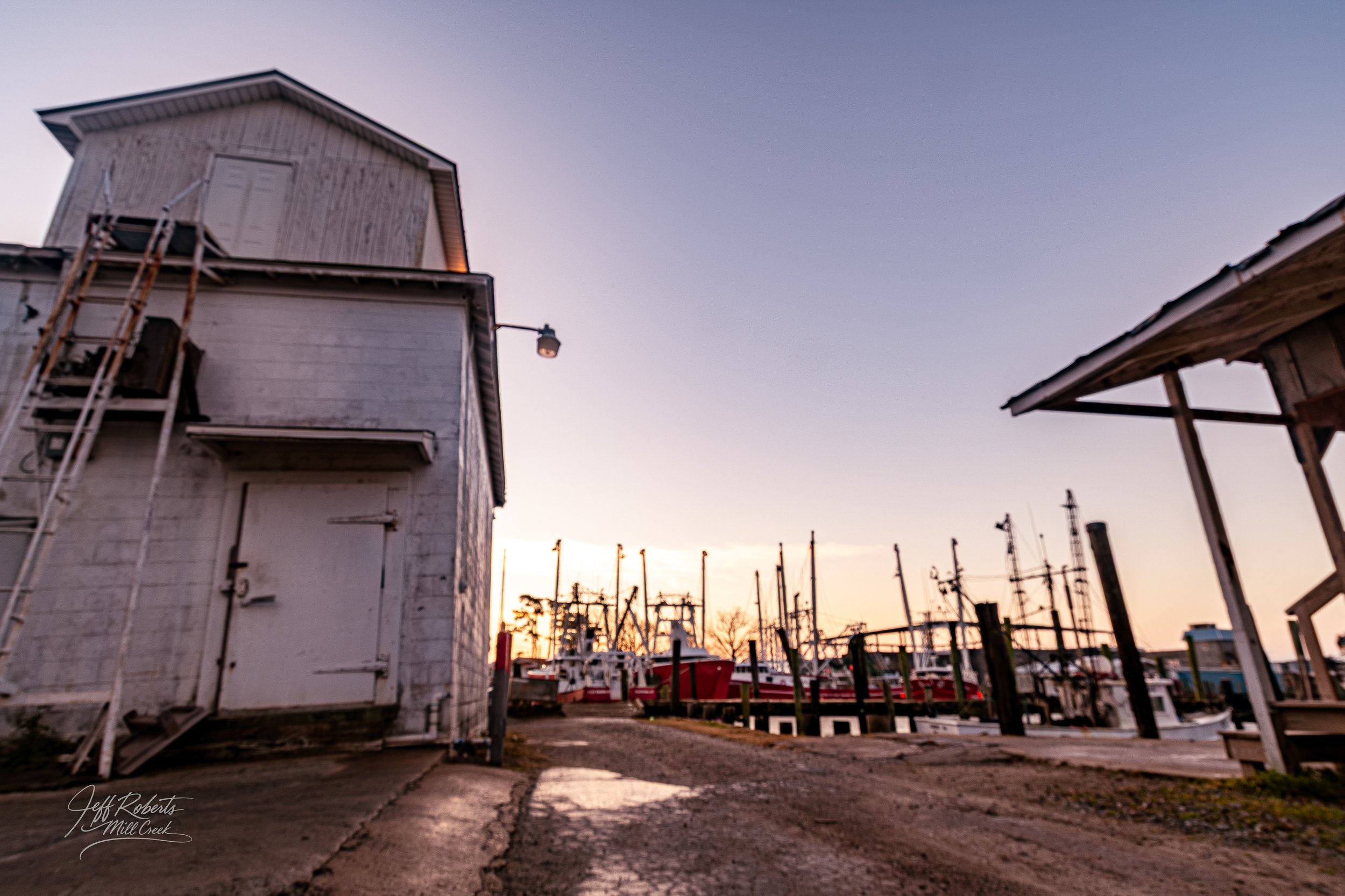 View of a marina at sunset with boats docked, a white wooden building on the left, and a small wooden structure on the right.