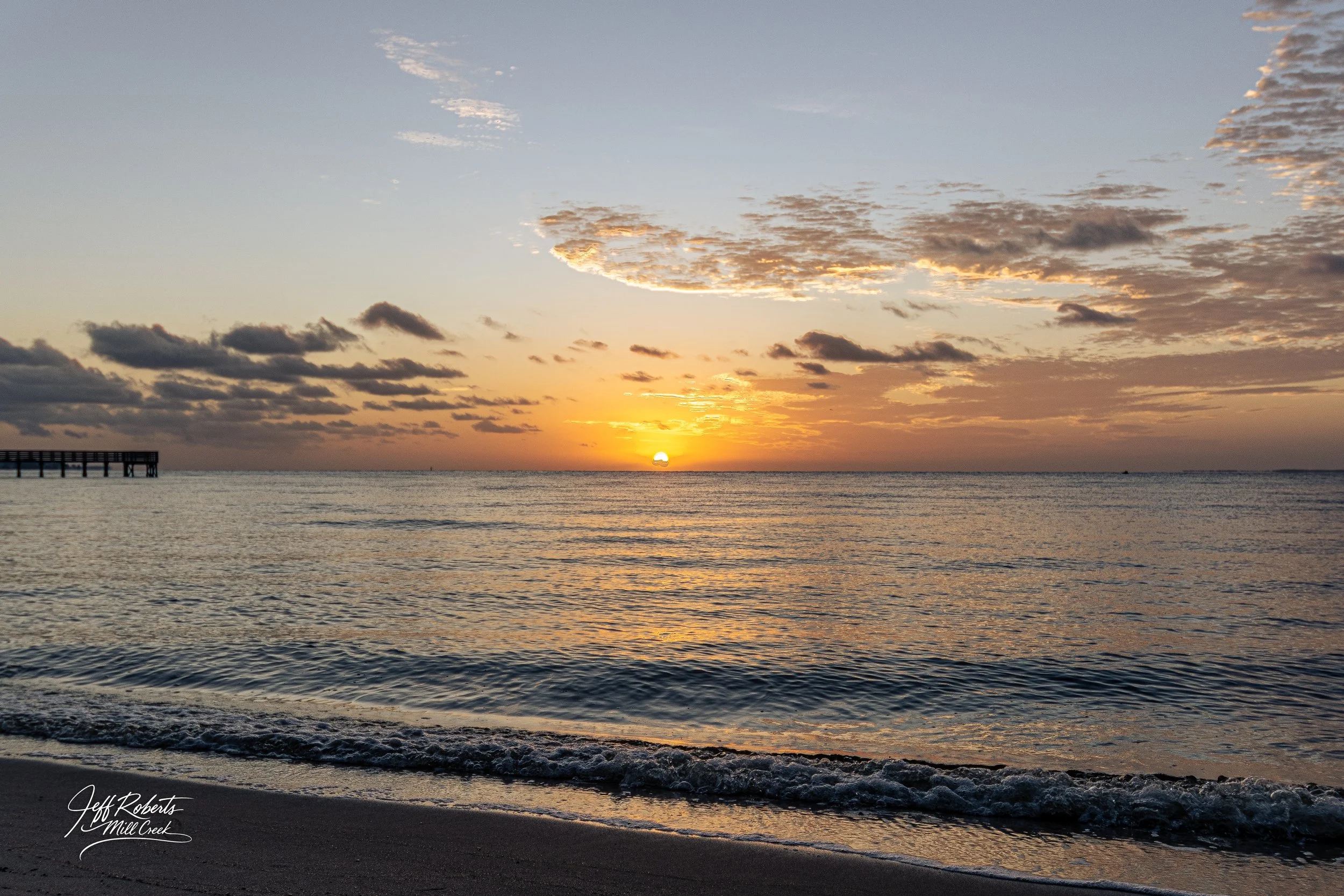 Sunset over the ocean with partly cloudy sky, gentle waves on the beach, and a pier on the left side.
