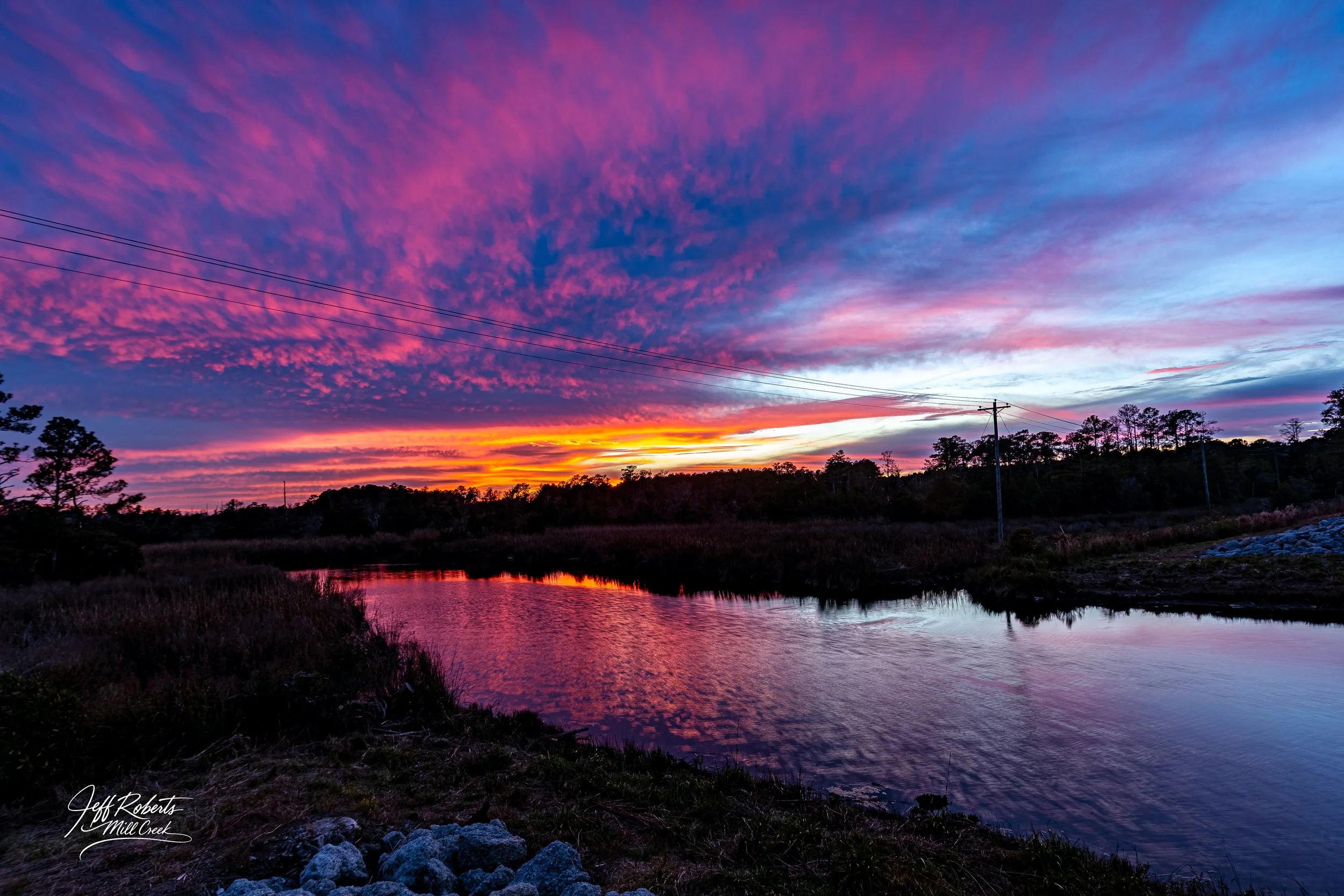 Colorful sunset over a river with pink, purple, and orange clouds, trees, and power lines in silhouette