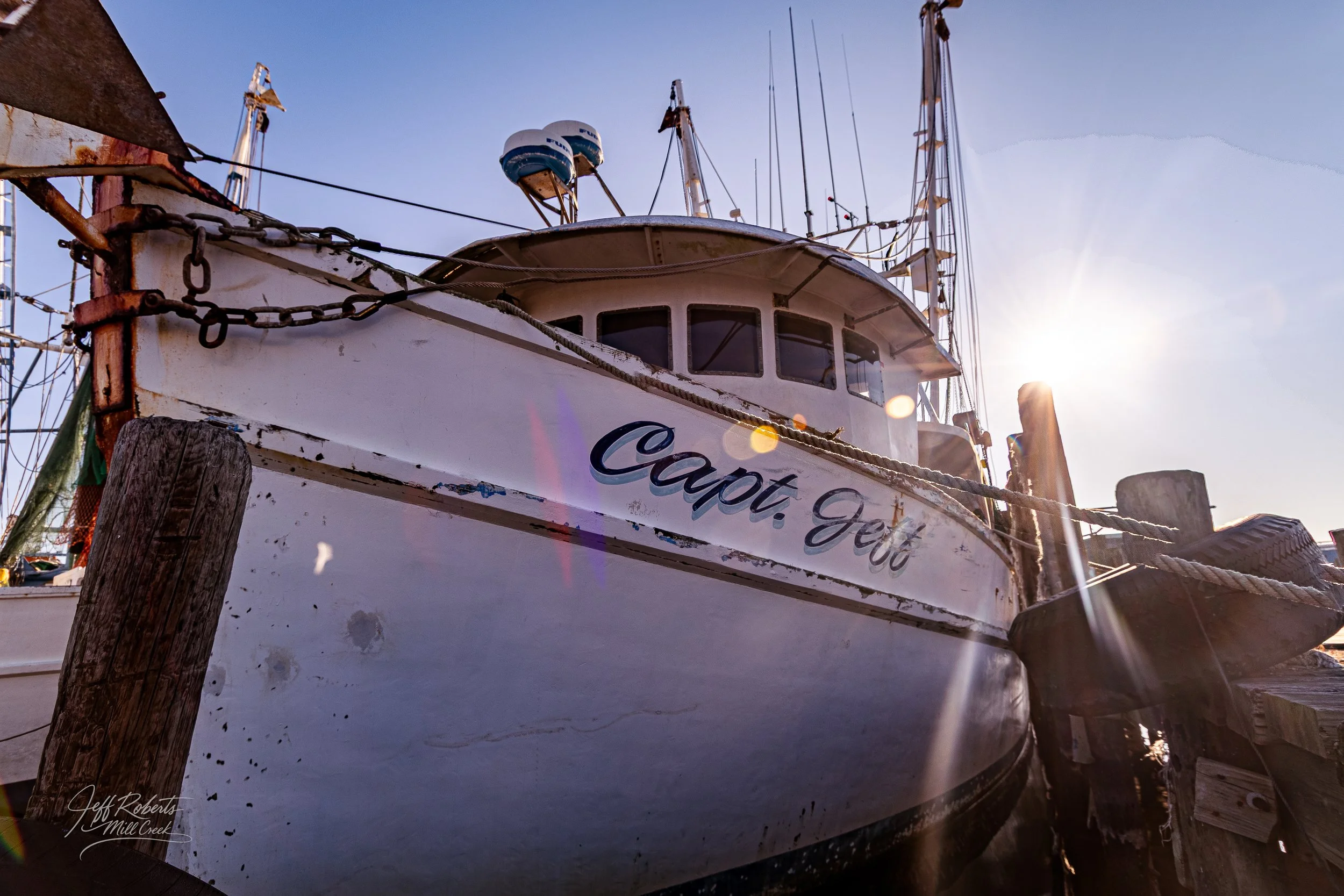 A white boat named 'Capt. Jeff' docked at a marina with the sun behind it and a clear sky, showing weathered paint and rigging equipment.