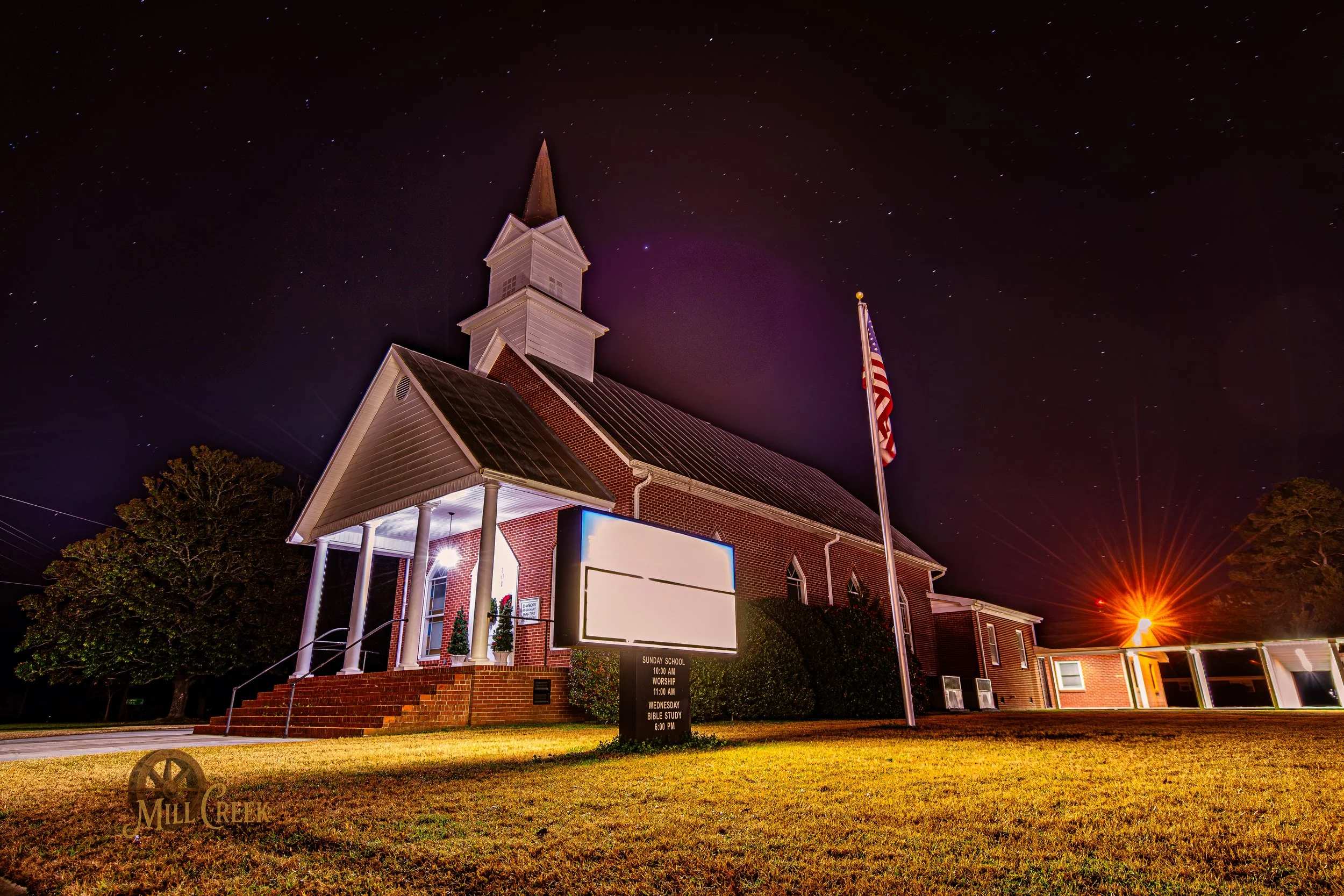 Nighttime view of a brick church with a tall steeple and an American flag in front. The church has a covered porch with stairs, illuminated by exterior lights. There is a sign in front of the church displaying service times and activities. The night 