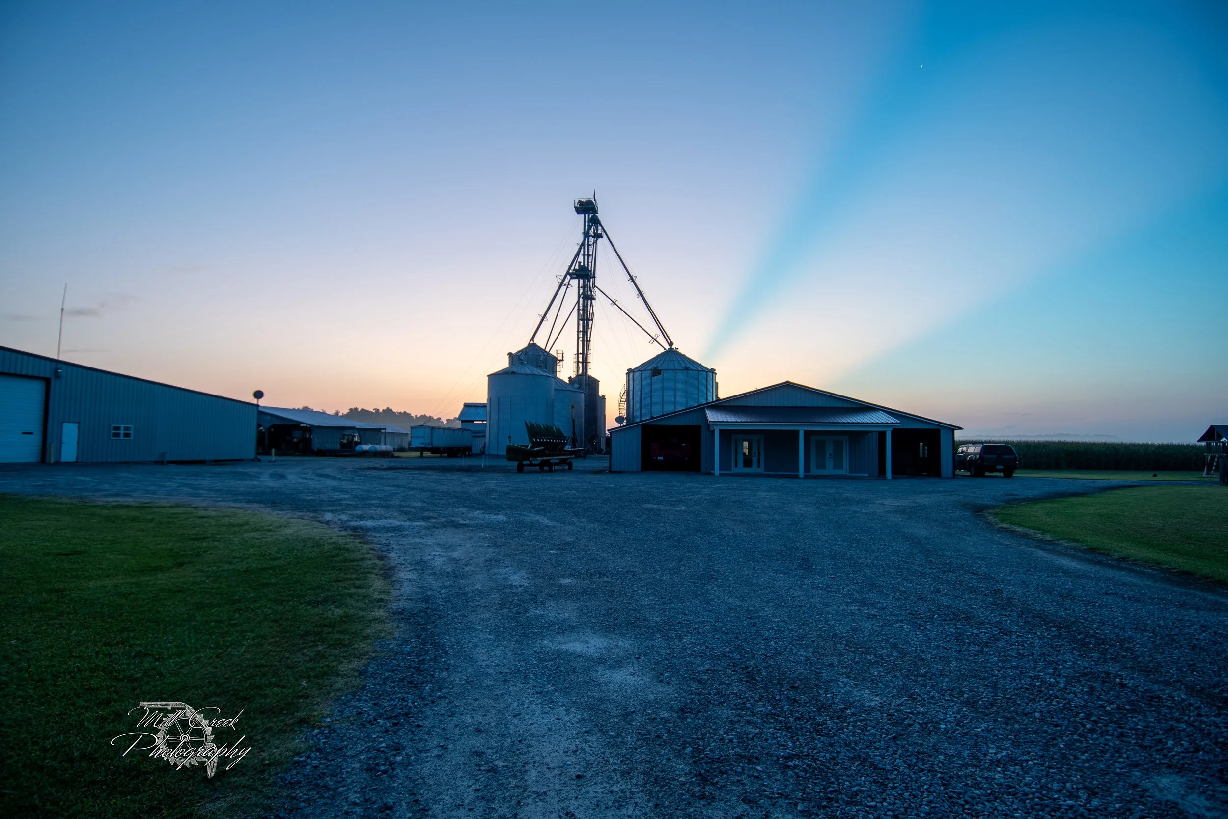 Silos at sunrise — Grain silos and farm buildings stand tall in silhouette, backlit by a soft pastel sunrise, with beams of light streaking across the sky like spotlights.