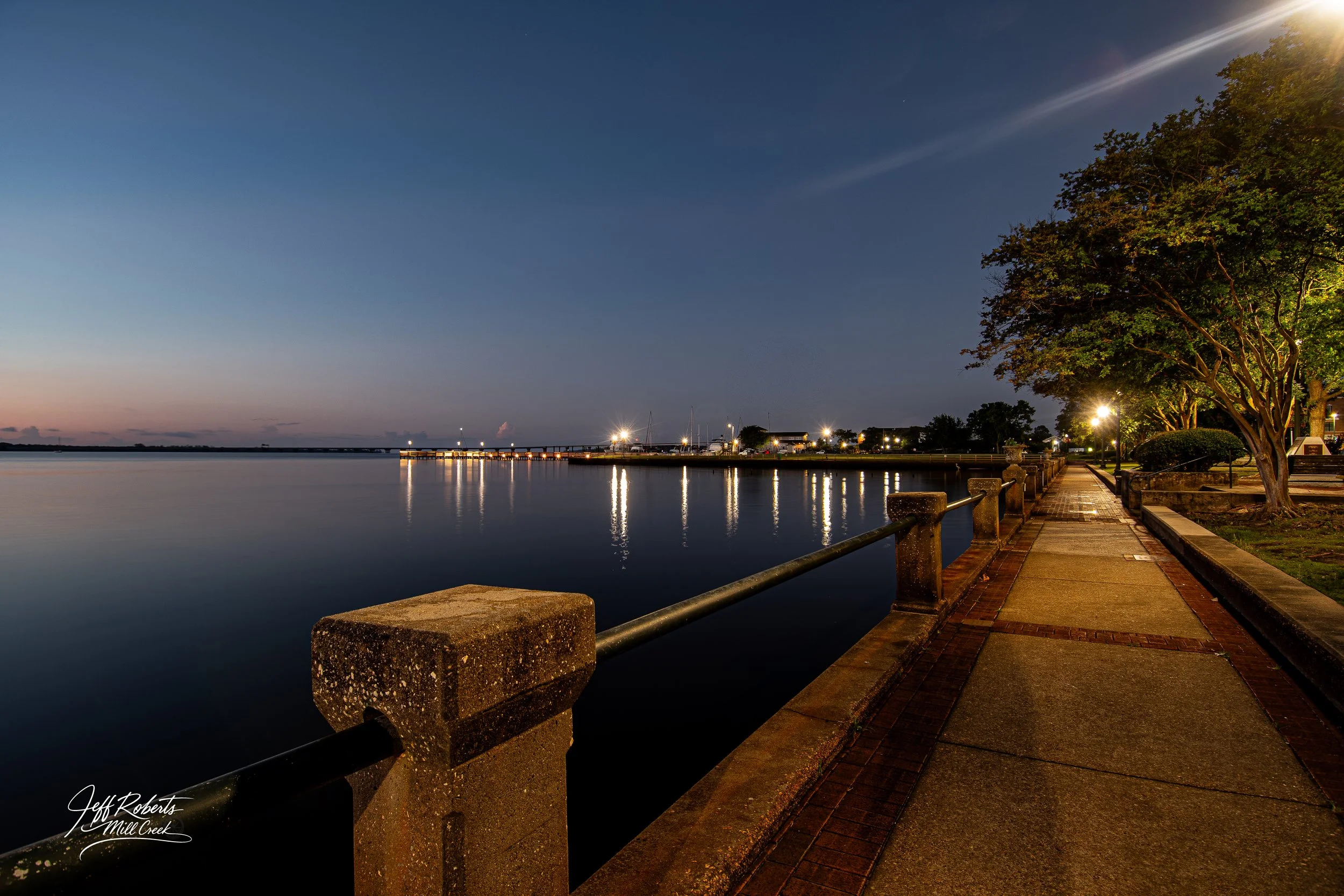 A peaceful waterfront promenade at dusk with a sidewalk, a railing along the water, trees illuminated by streetlights, and a calm body of water reflecting city lights.