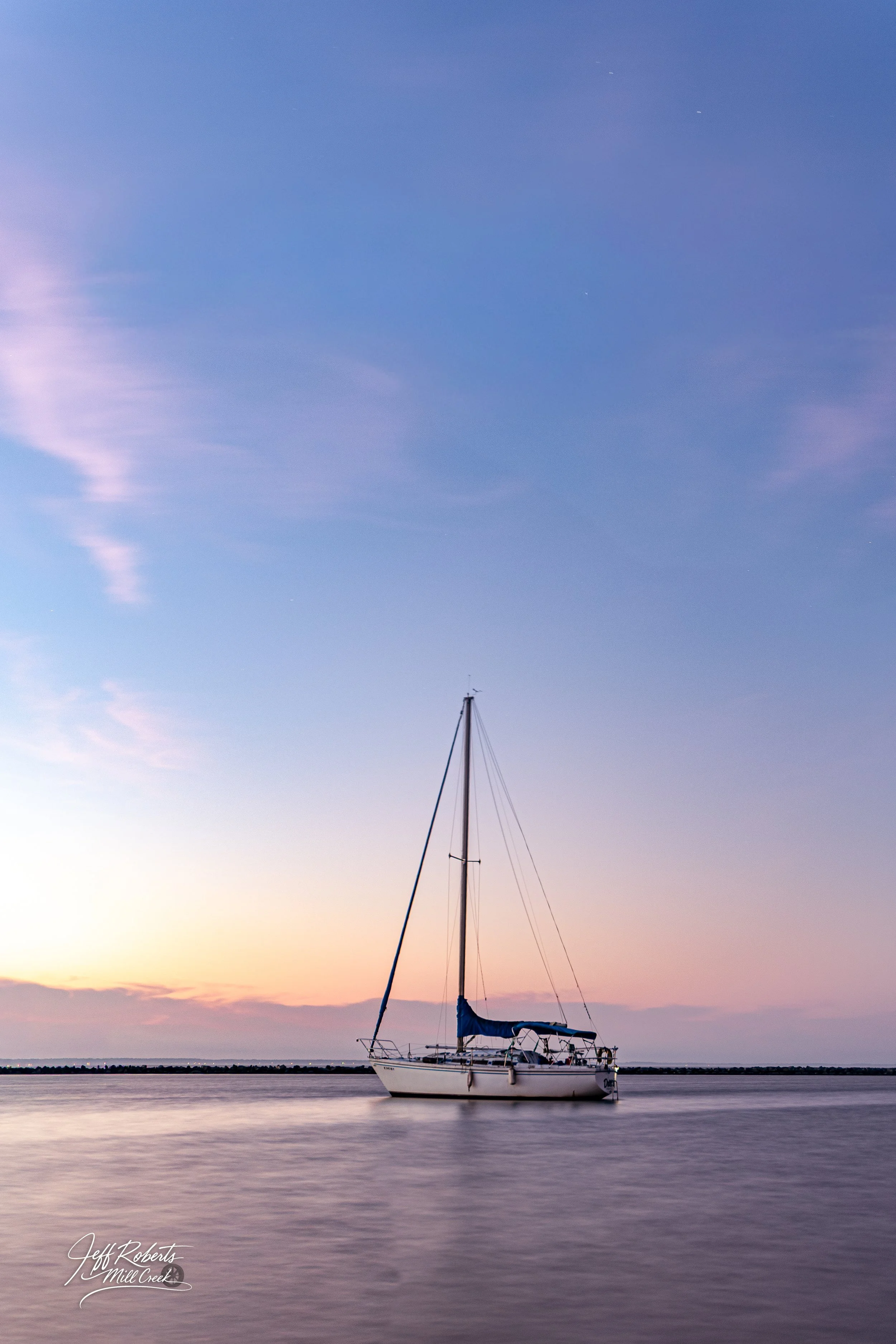 A sailboat floating on calm water during sunset or sunrise, with a mostly clear sky and some light clouds.