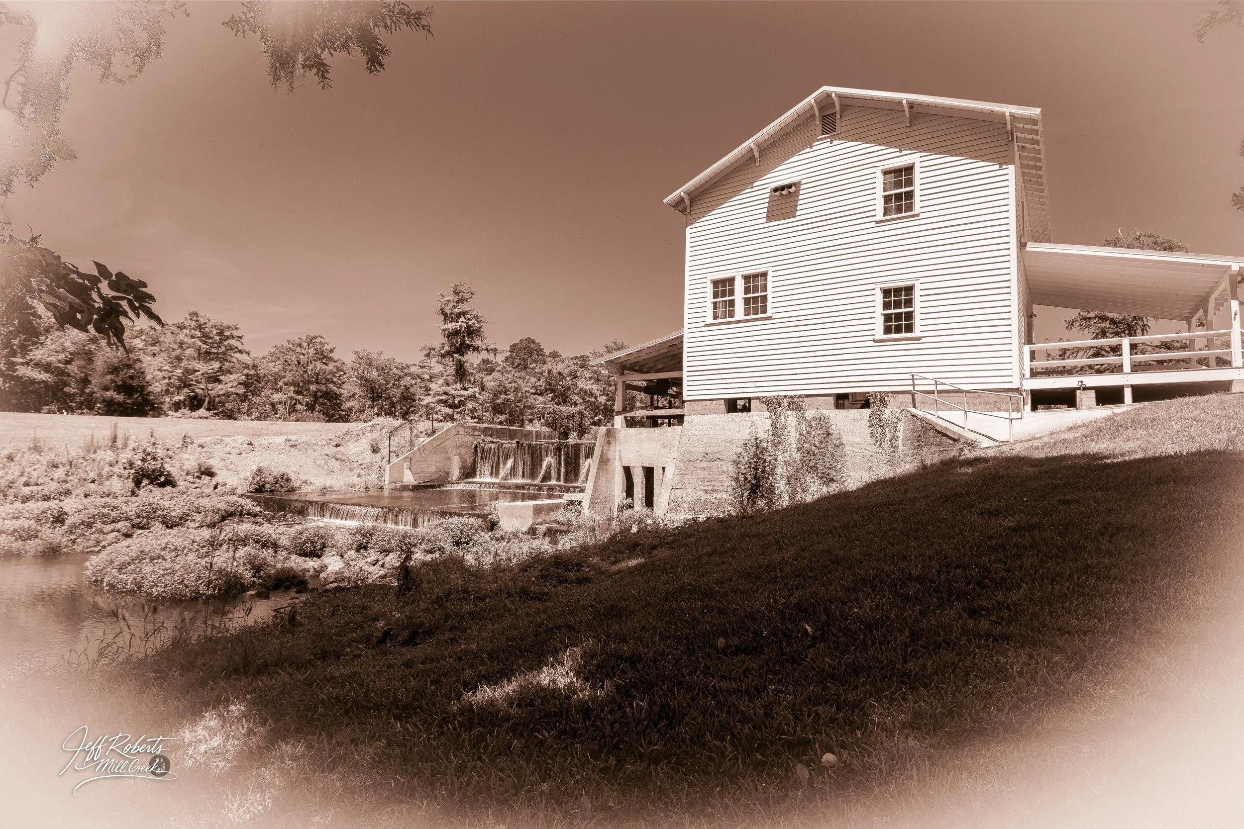 Sepia-toned photo of a two-story white house with a steep roof, located on a grassy hill near a small waterfall and pond, with trees in the background.