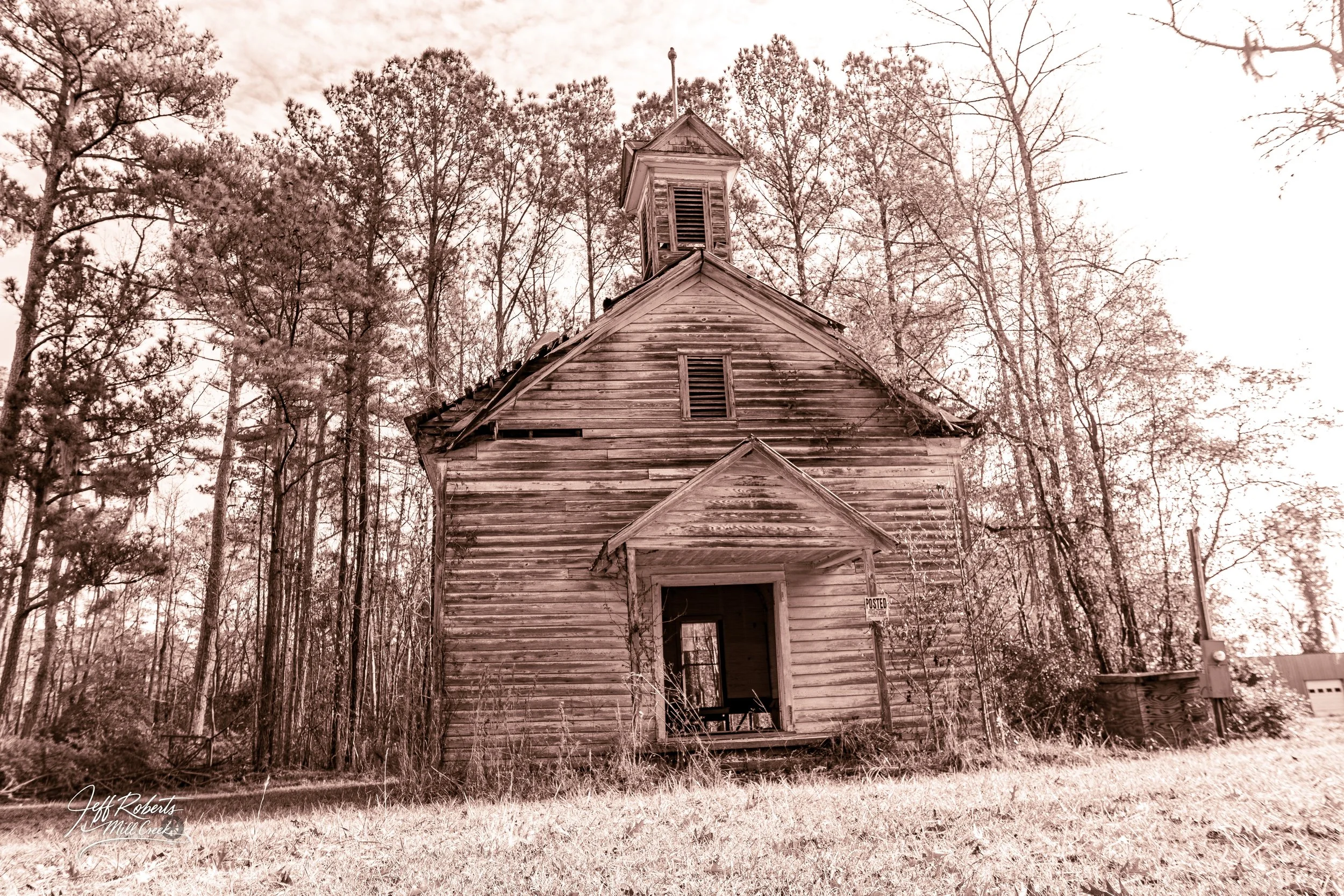 An abandoned, weathered wooden church surrounded by tall, leafless trees, with a sign that reads 'POSTED' near the entrance, set against a cloudy sky.