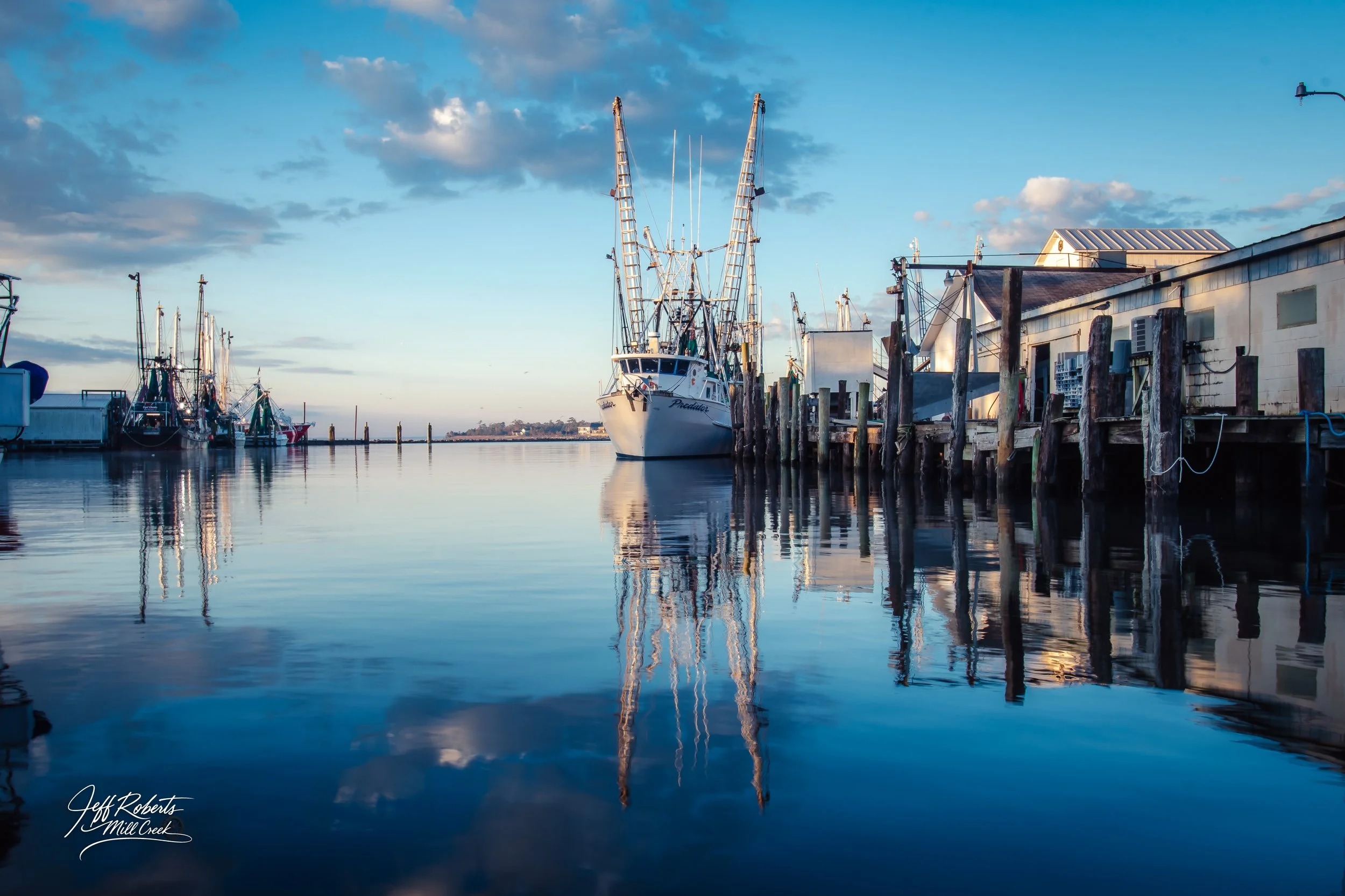 Boats docked at a marina with calm water reflecting the boats and sky, with wooden piers and a building on the right side.