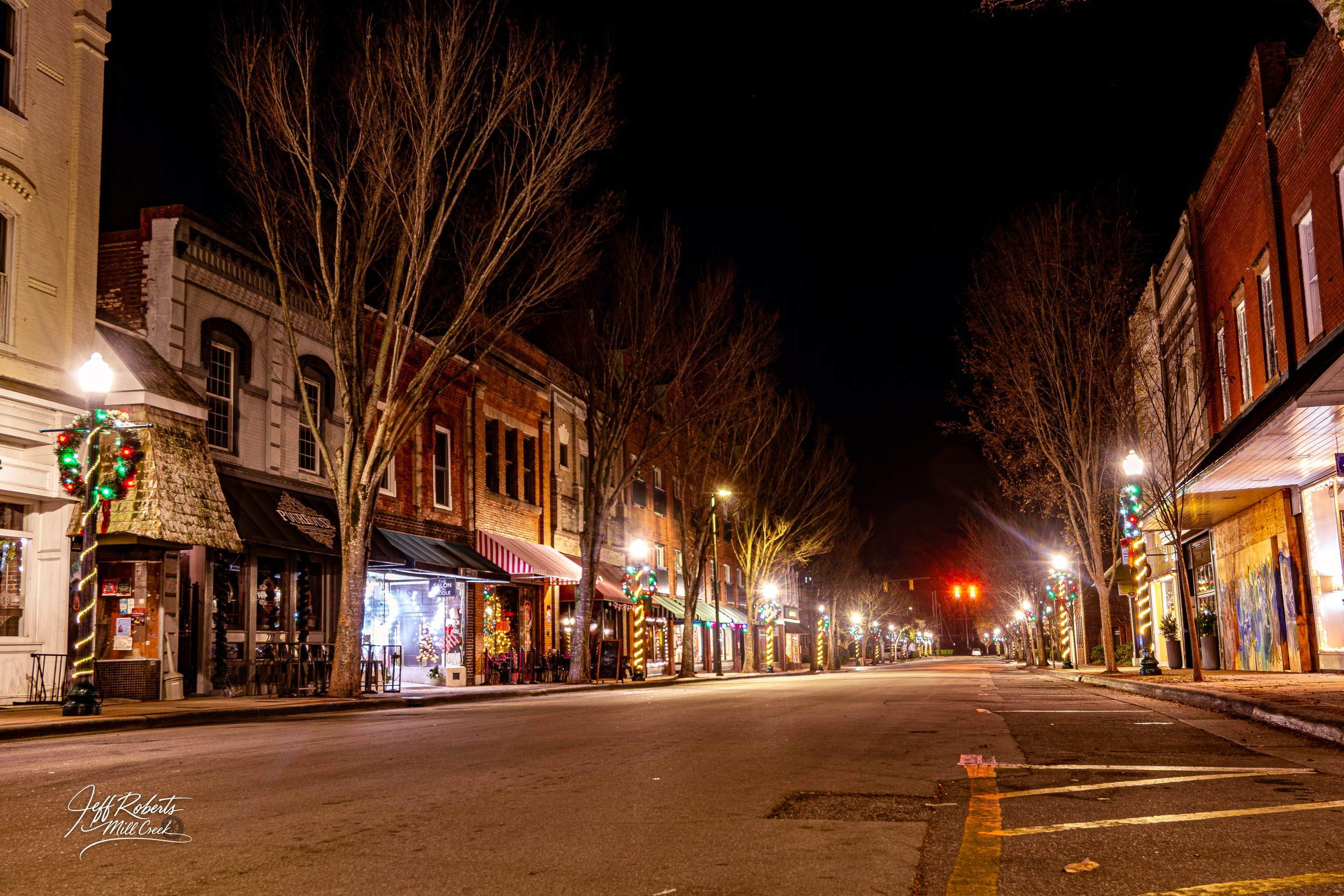 Empty downtown street at night decorated with Christmas lights on lamp posts and storefronts, with bare trees and a dark sky above.