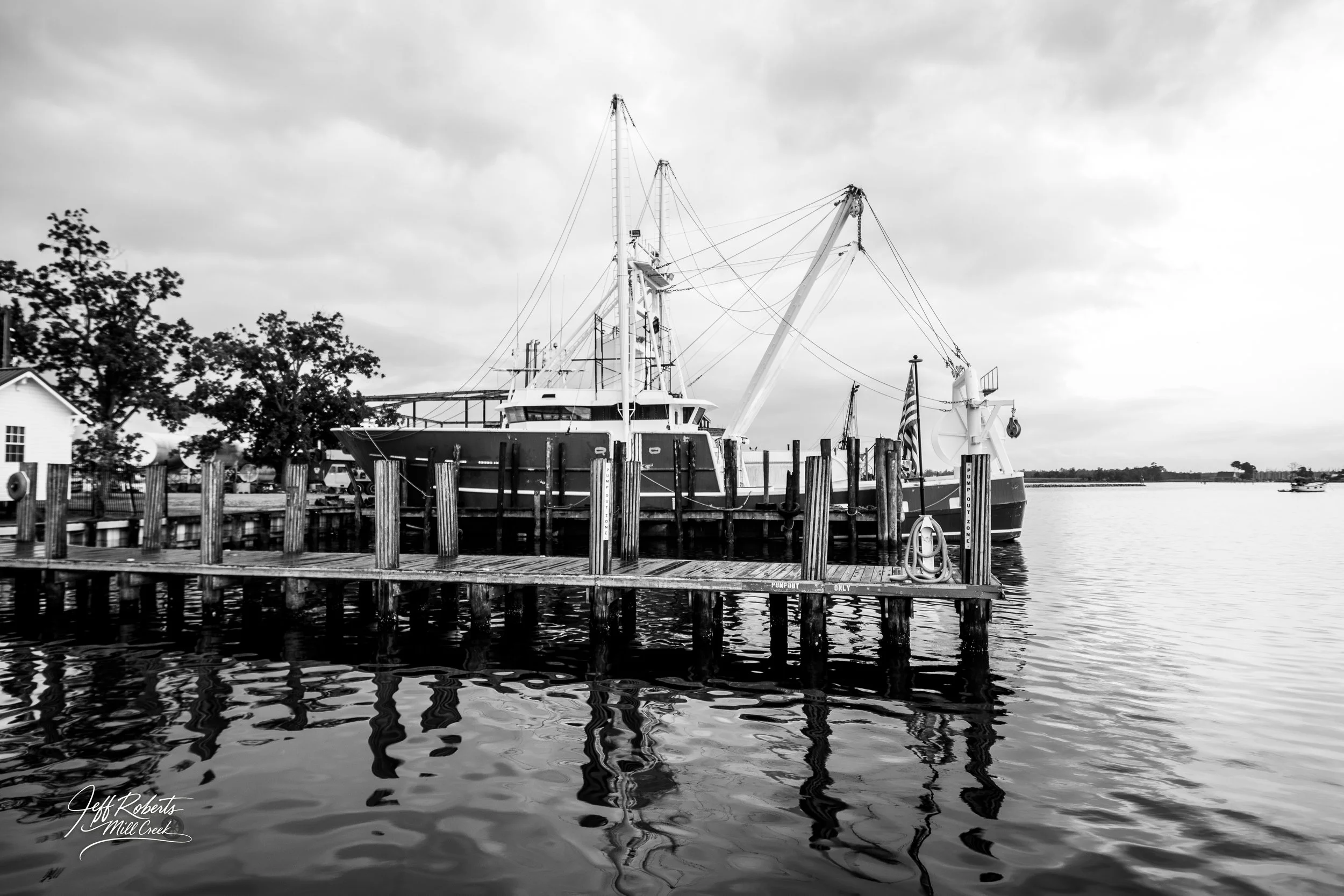 A black and white photo of a large fishing boat docked at a wooden pier on a body of water. The boat has tall masts and a crane, and there are trees and small buildings along the shoreline in the background.