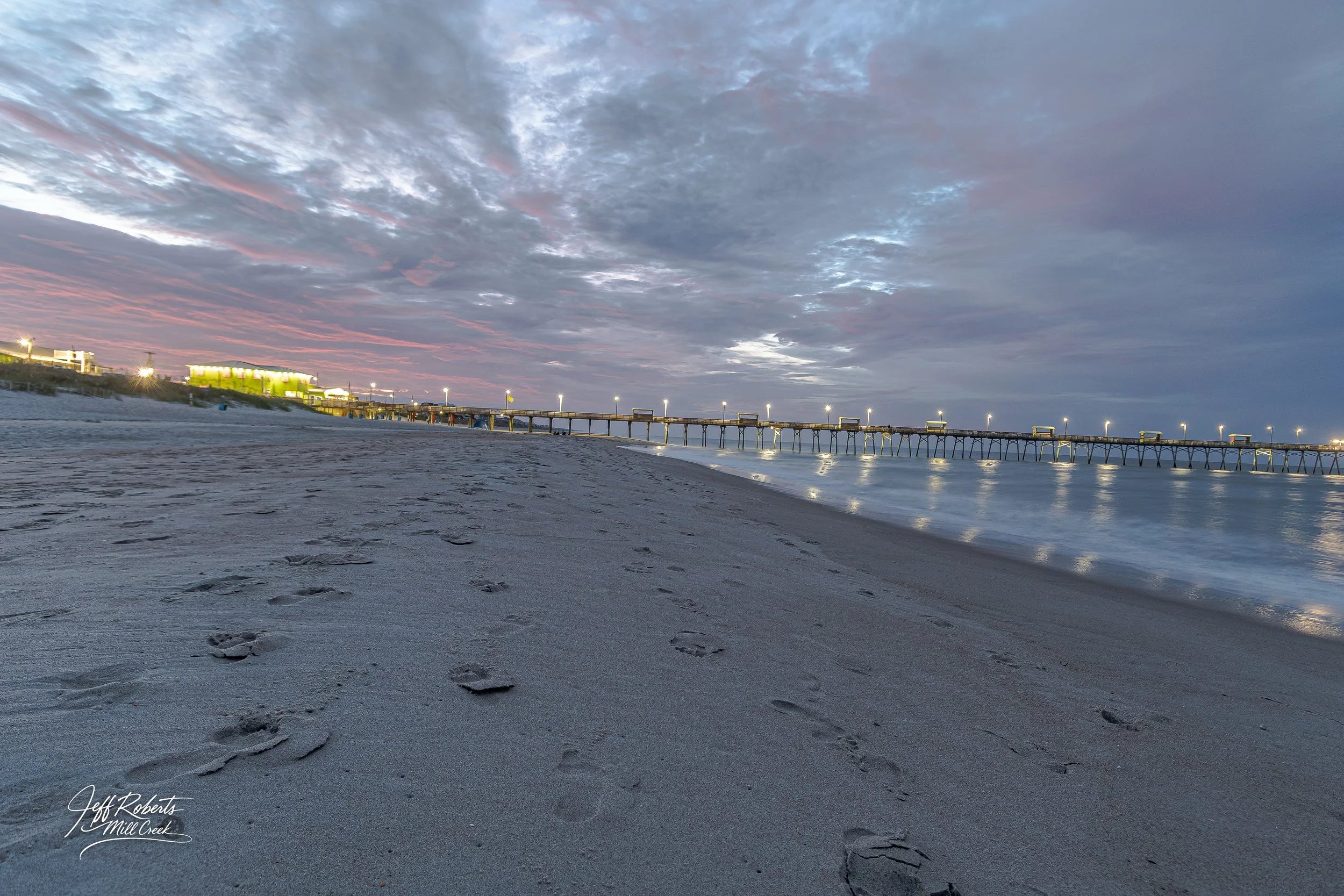 A sandy beach with footprints leading toward a pier extending over calm water during sunset or sunrise, with a partly cloudy sky and some buildings lit up in the distance.