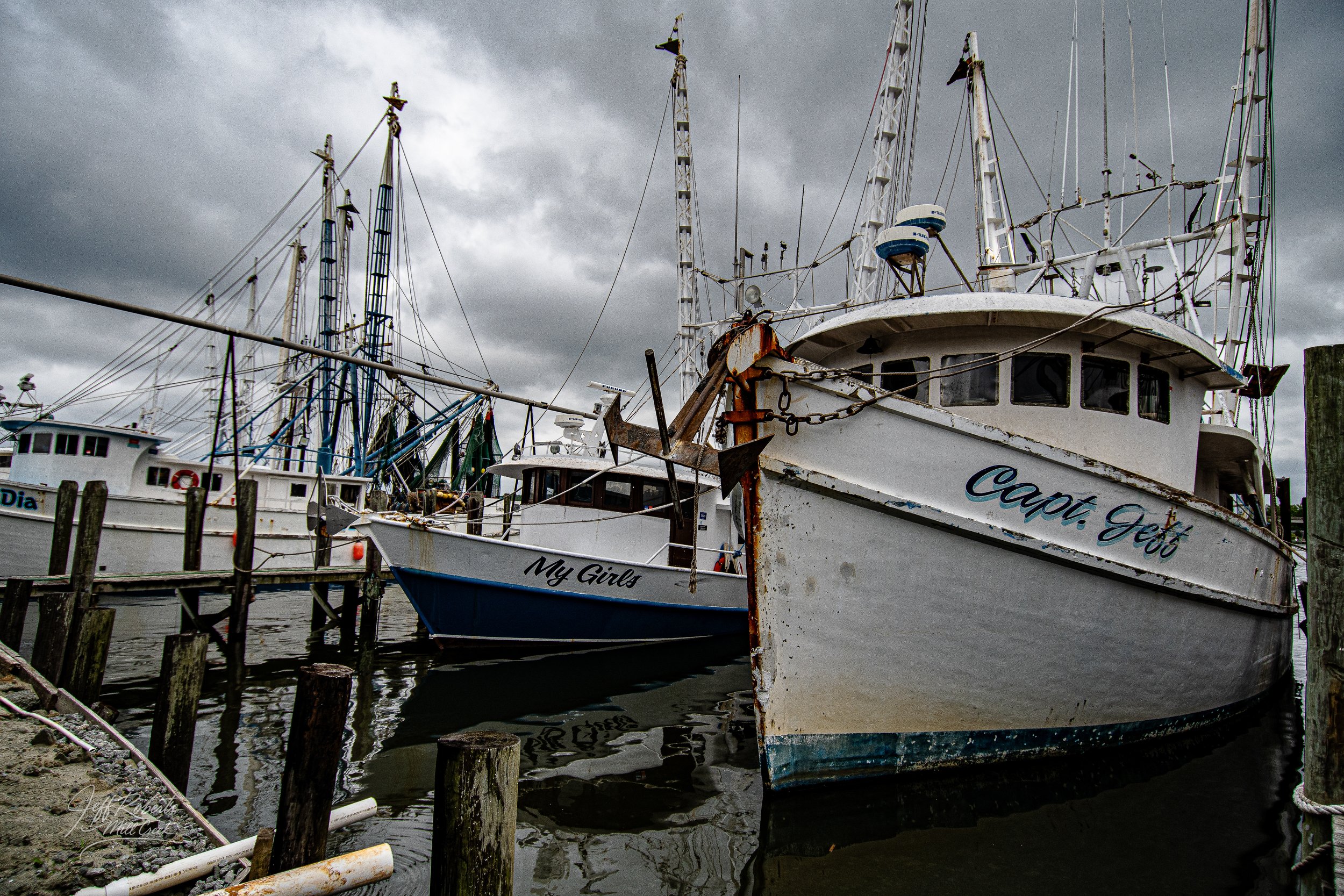 Several boats docked at a marina under dark, cloudy skies, including one named 'Capit. Jeff' and another named 'My Girls', with weathered wood pilings and water reflections.