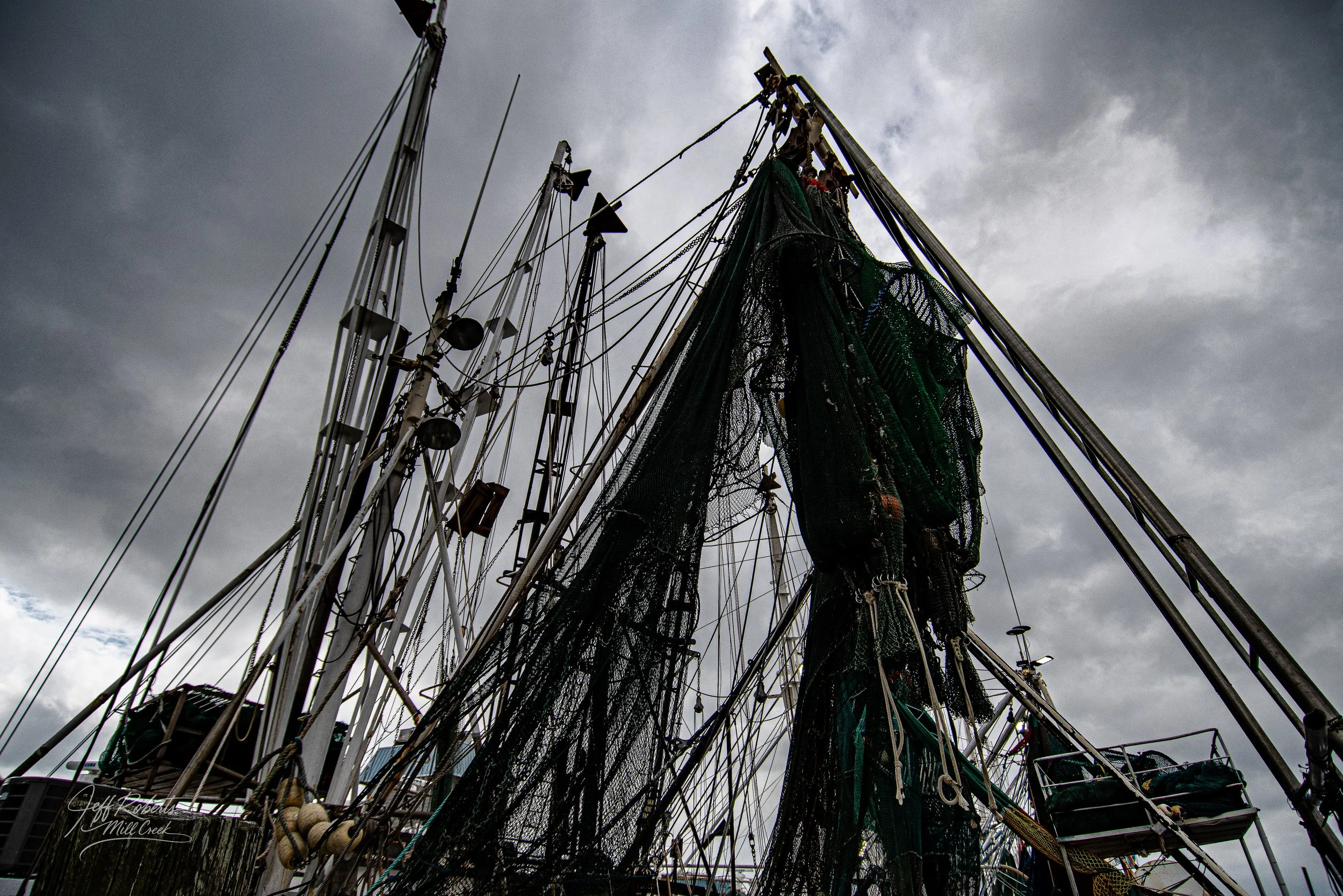 View of fishing boat masts and rigging against a cloudy sky, with fishing nets hanging from the boat.