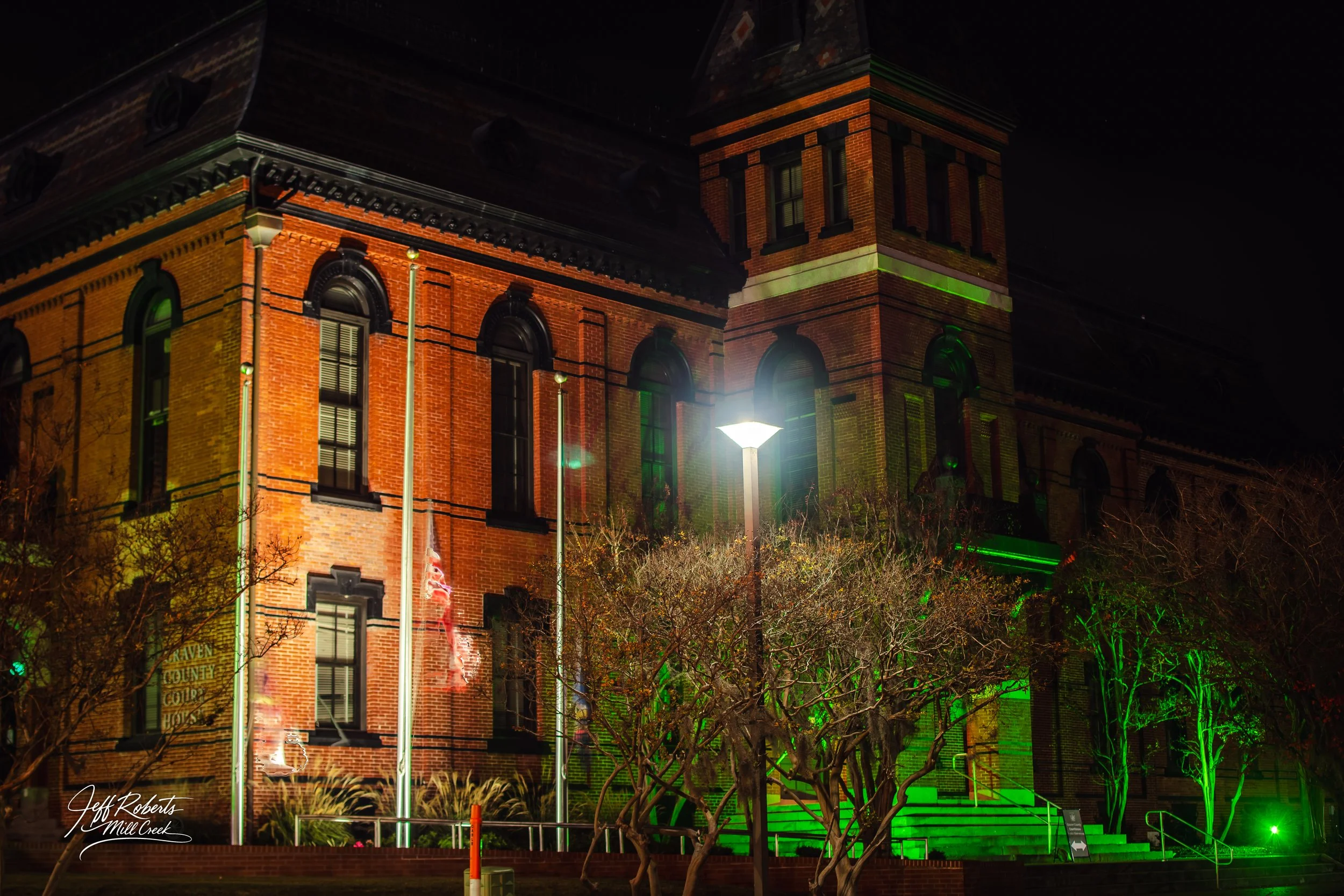 Nighttime view of a historic brick building illuminated with green and white lights, with steps leading up to the entrance and leafless trees in front.