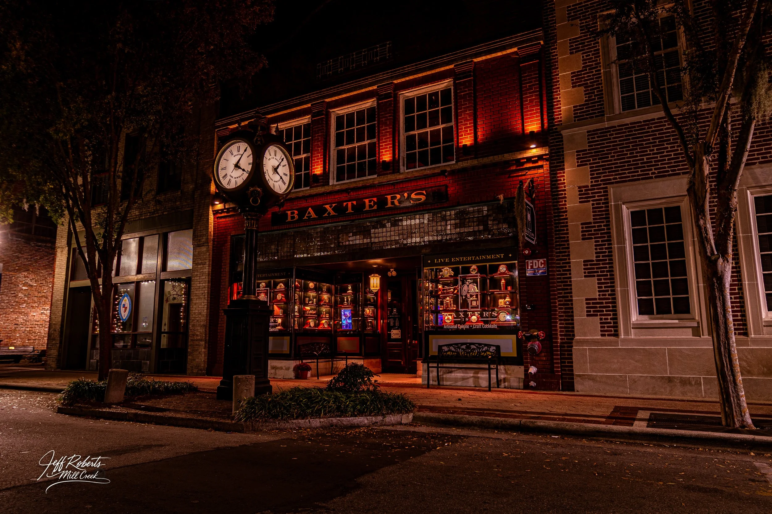 Nighttime view of Baxter's restaurant with illuminated red signage, lit display window, a street clock showing approximately 8:02, and trees lining the sidewalk.