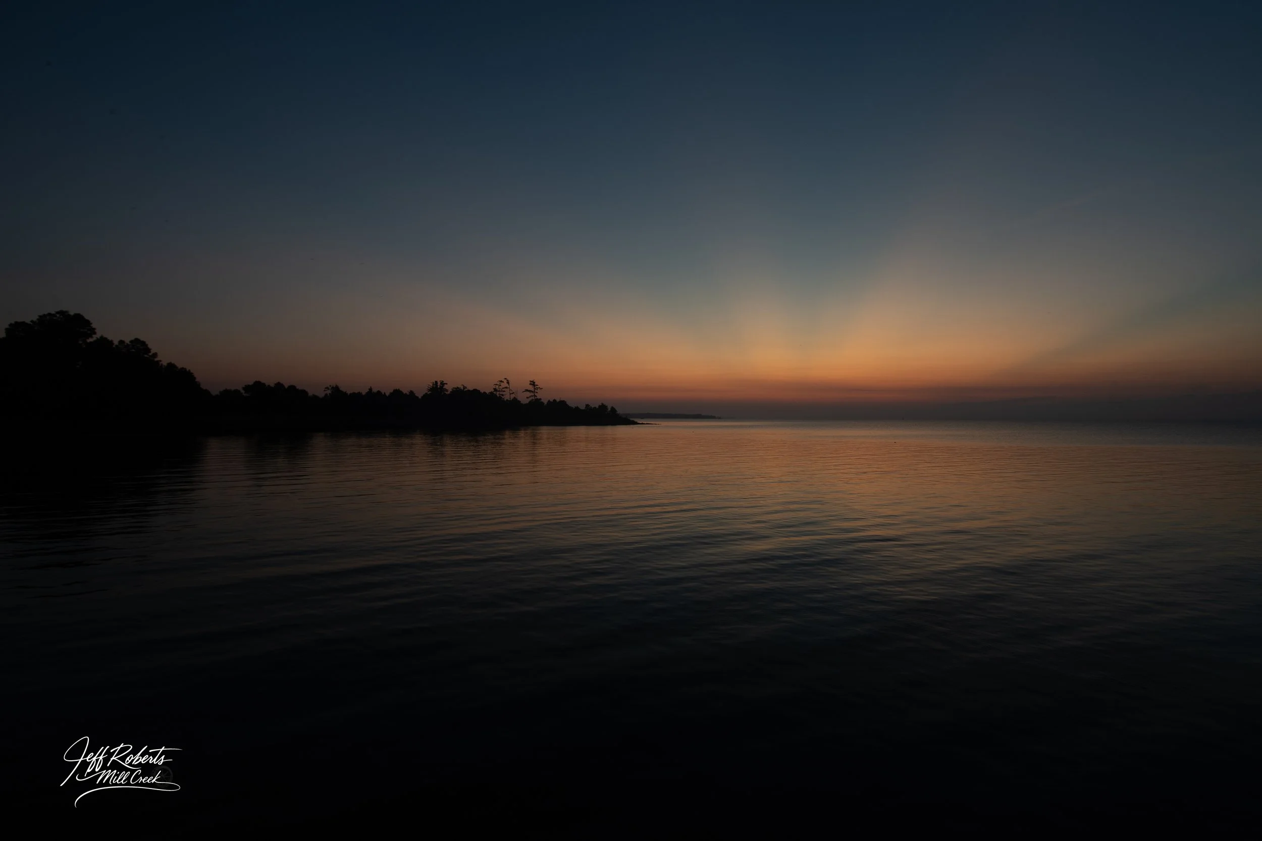 Sunset over a calm body of water with a silhouetted shoreline and trees in the distance.