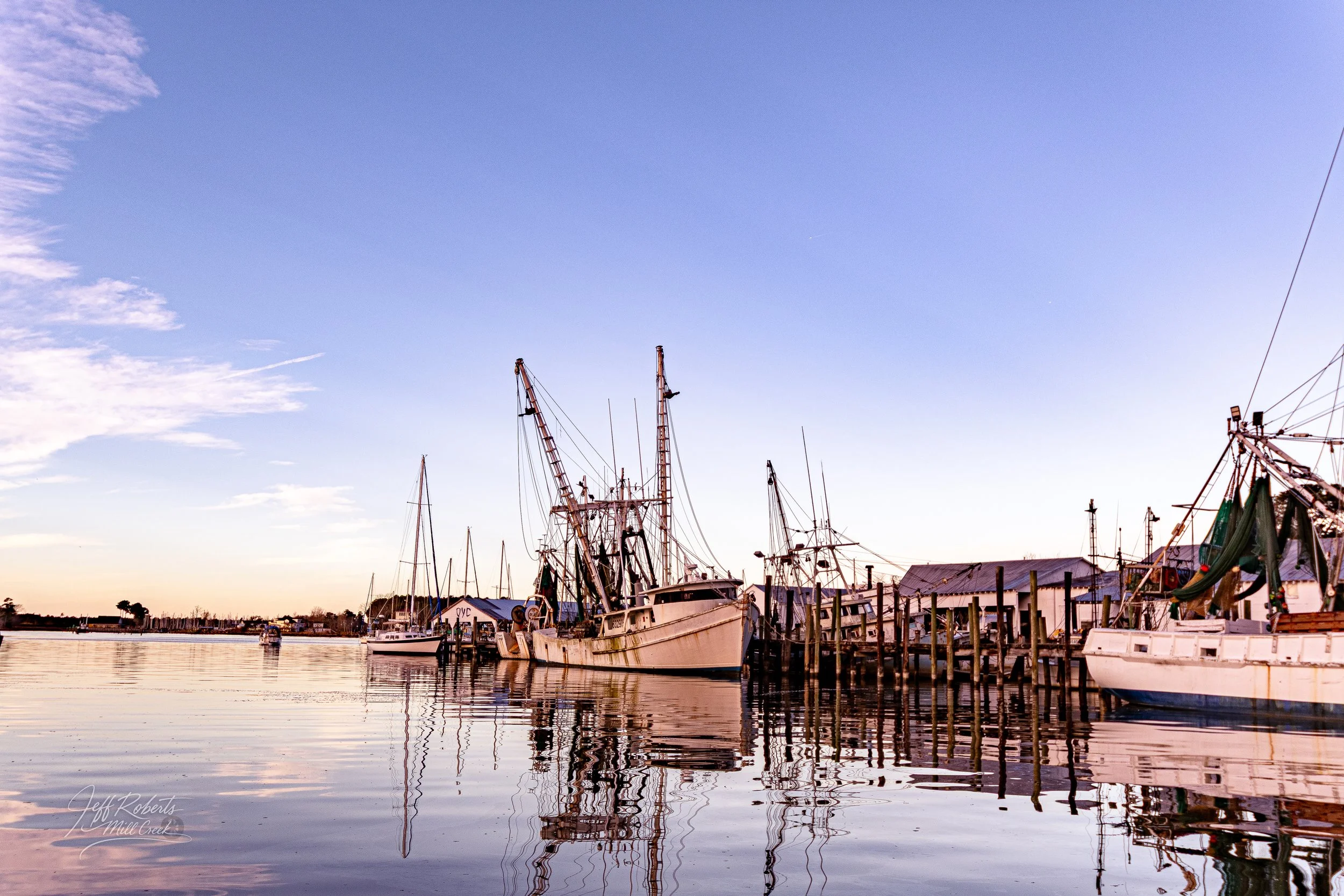Boats docked at a marina during sunset, with calm water reflecting the boats and a clear, blue sky.