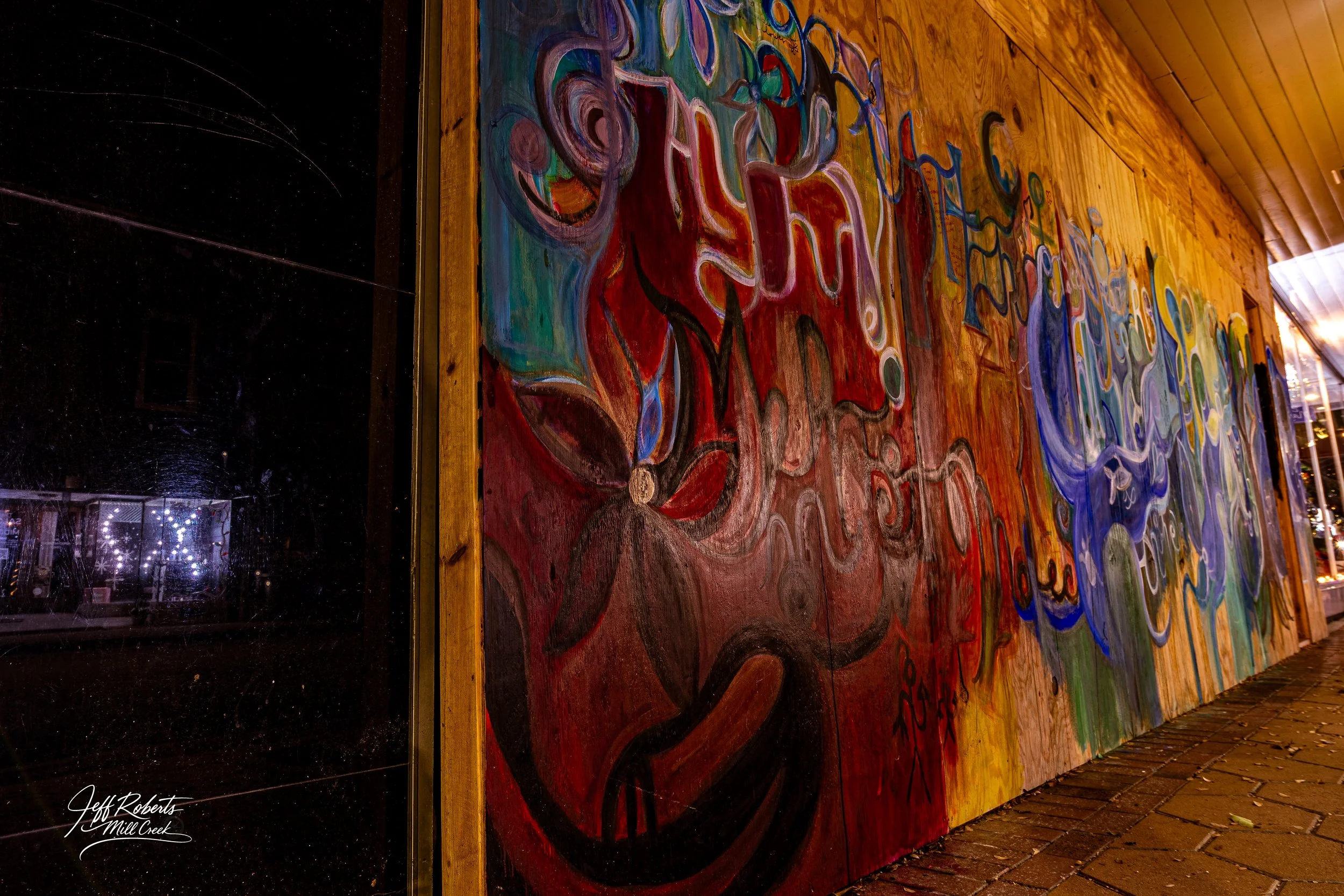 Colorful abstract mural painted on a wooden wall, featuring swirling shapes and vibrant colors, located outside at night with a brick sidewalk in front.
