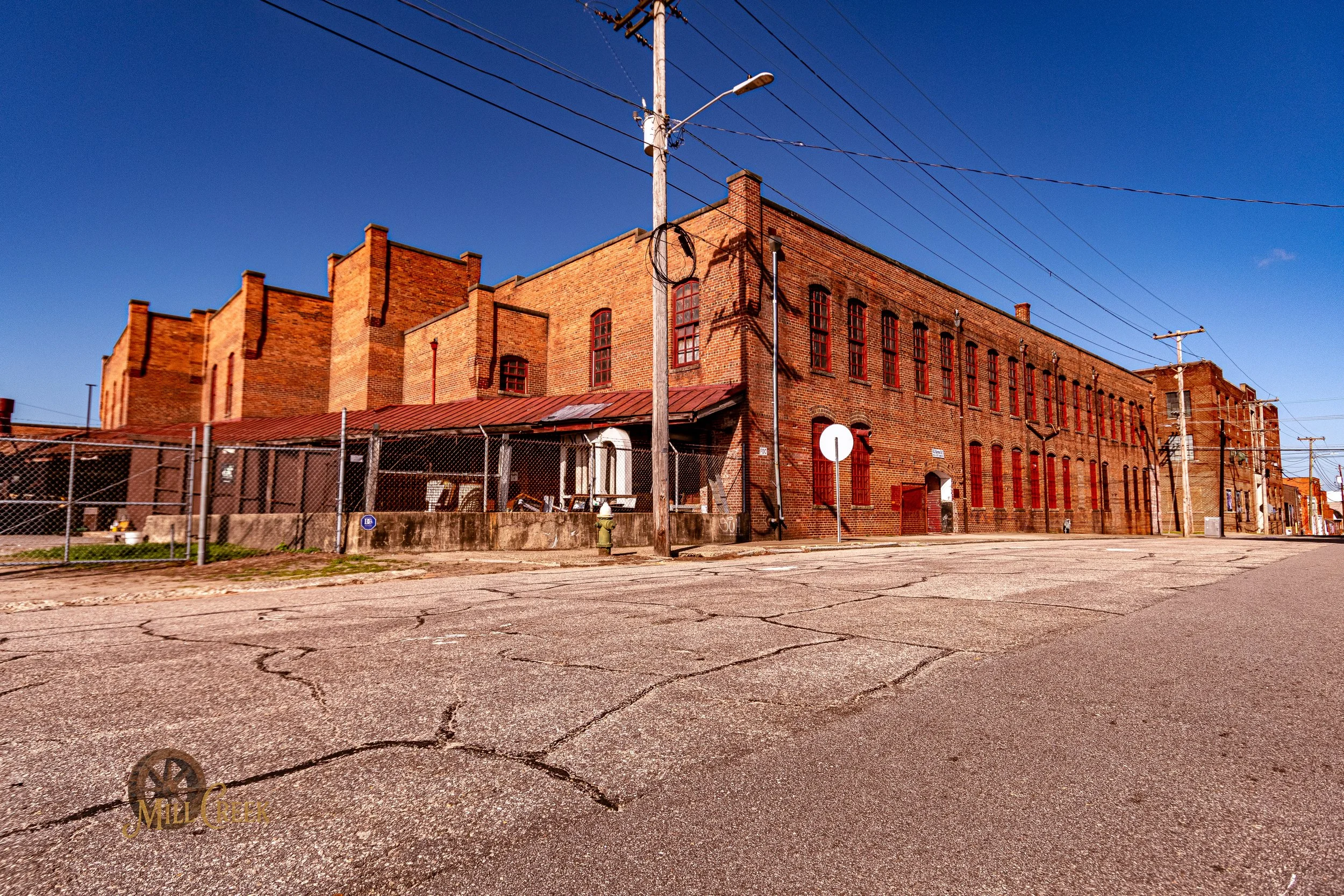 A large red brick industrial building with multiple windows, surrounded by a chain-link fence, under a clear blue sky.