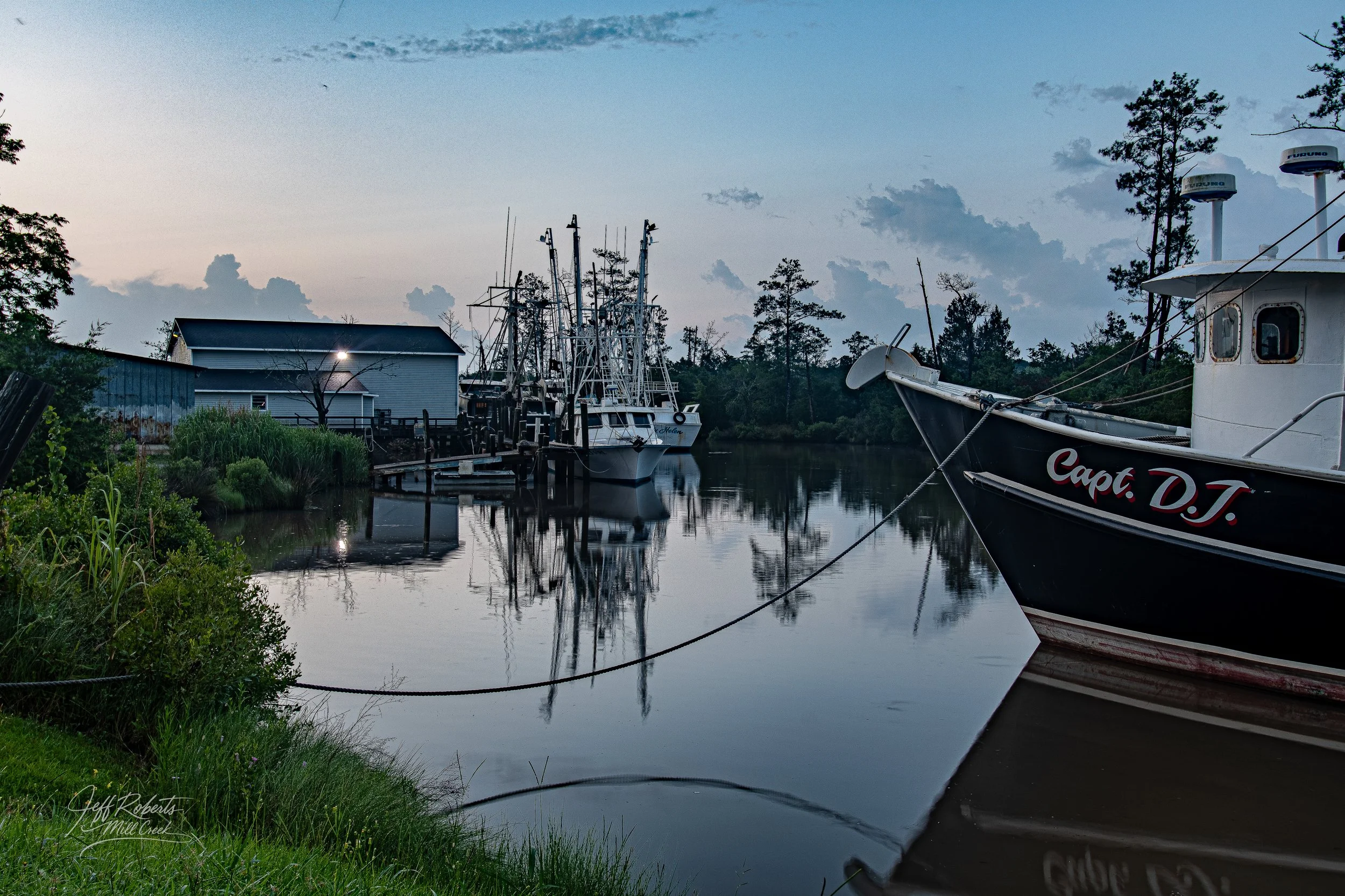 Boats docked along a calm river at dusk, with trees and a building in the background.