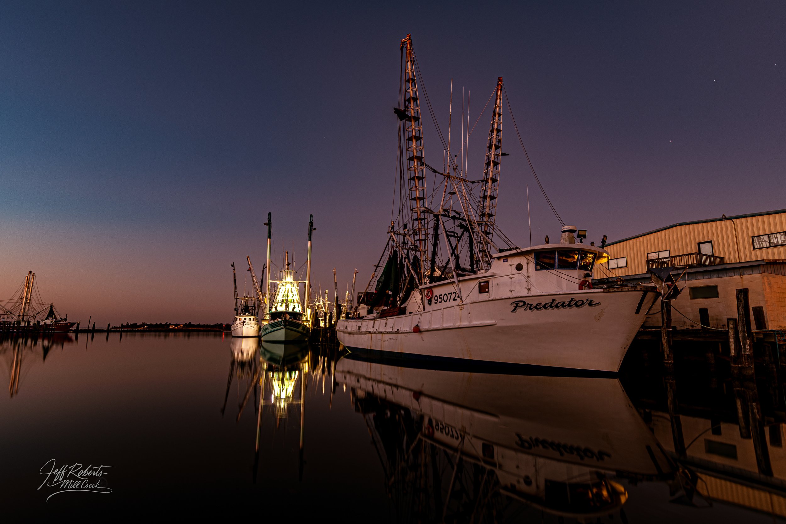 Boats docked on calm water at dusk with clear sky and buildings in the background.