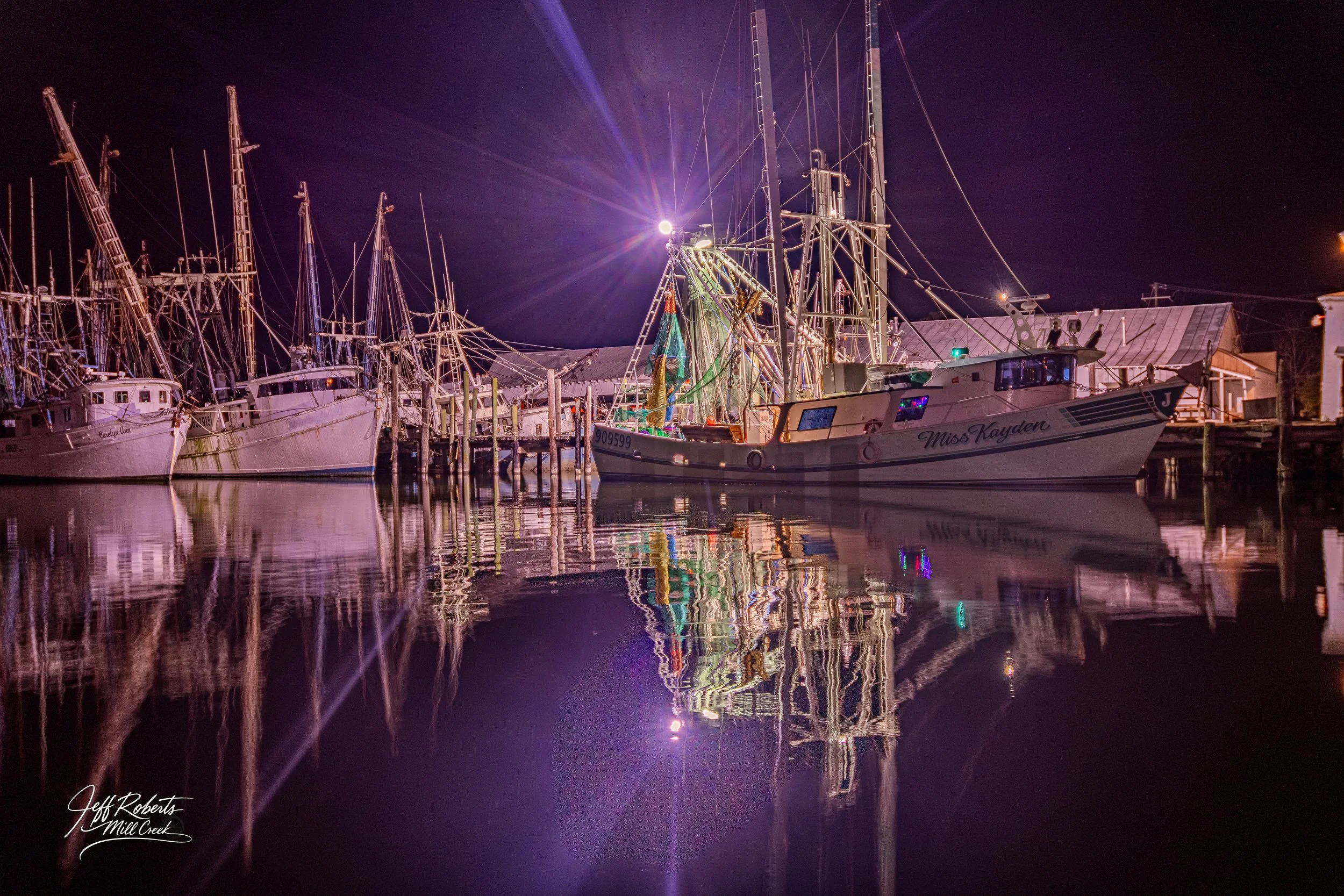 Nighttime scene at a marina with several boats docked, including one named Miss Kayden, illuminated by bright lights that reflect on the calm water, with the sky dark and a few stars visible.