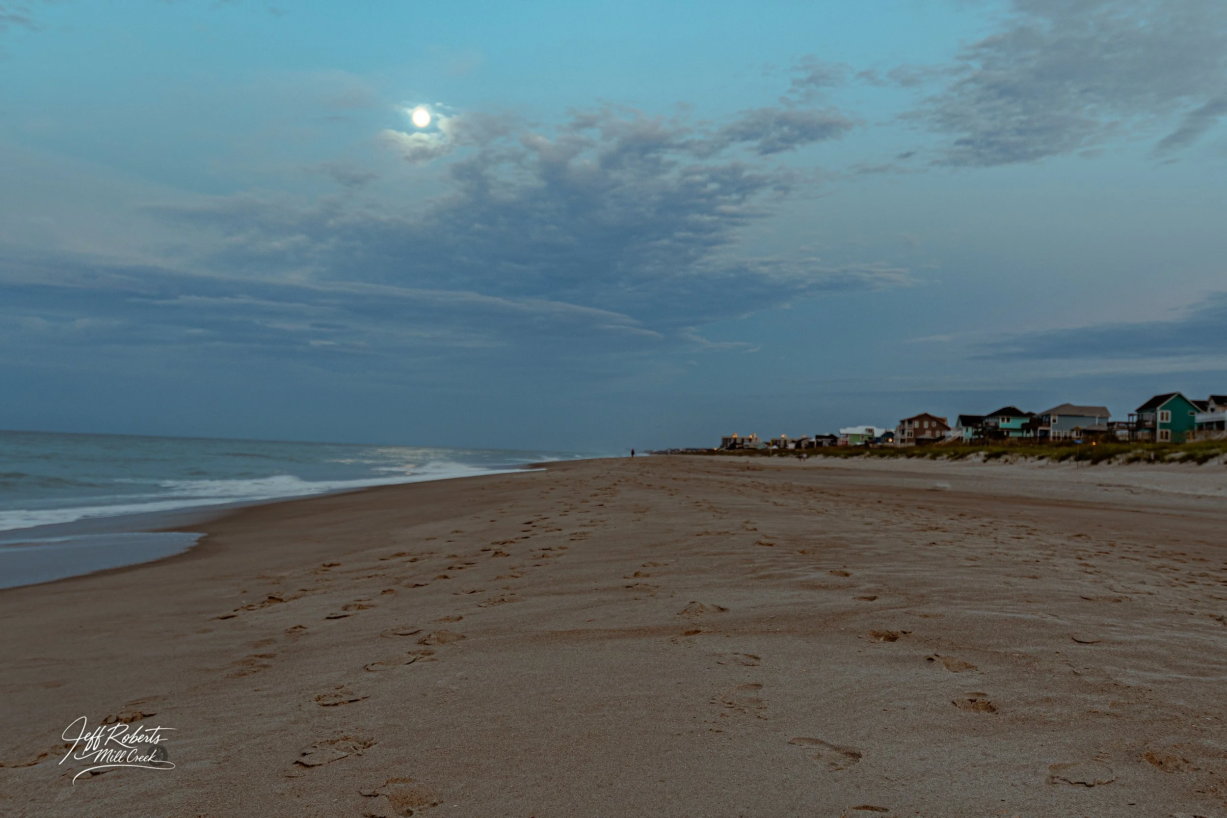 Empty beach with footprints in the sand, ocean waves on the left, houses in the distance on the right, cloudy sky with the moon visible.