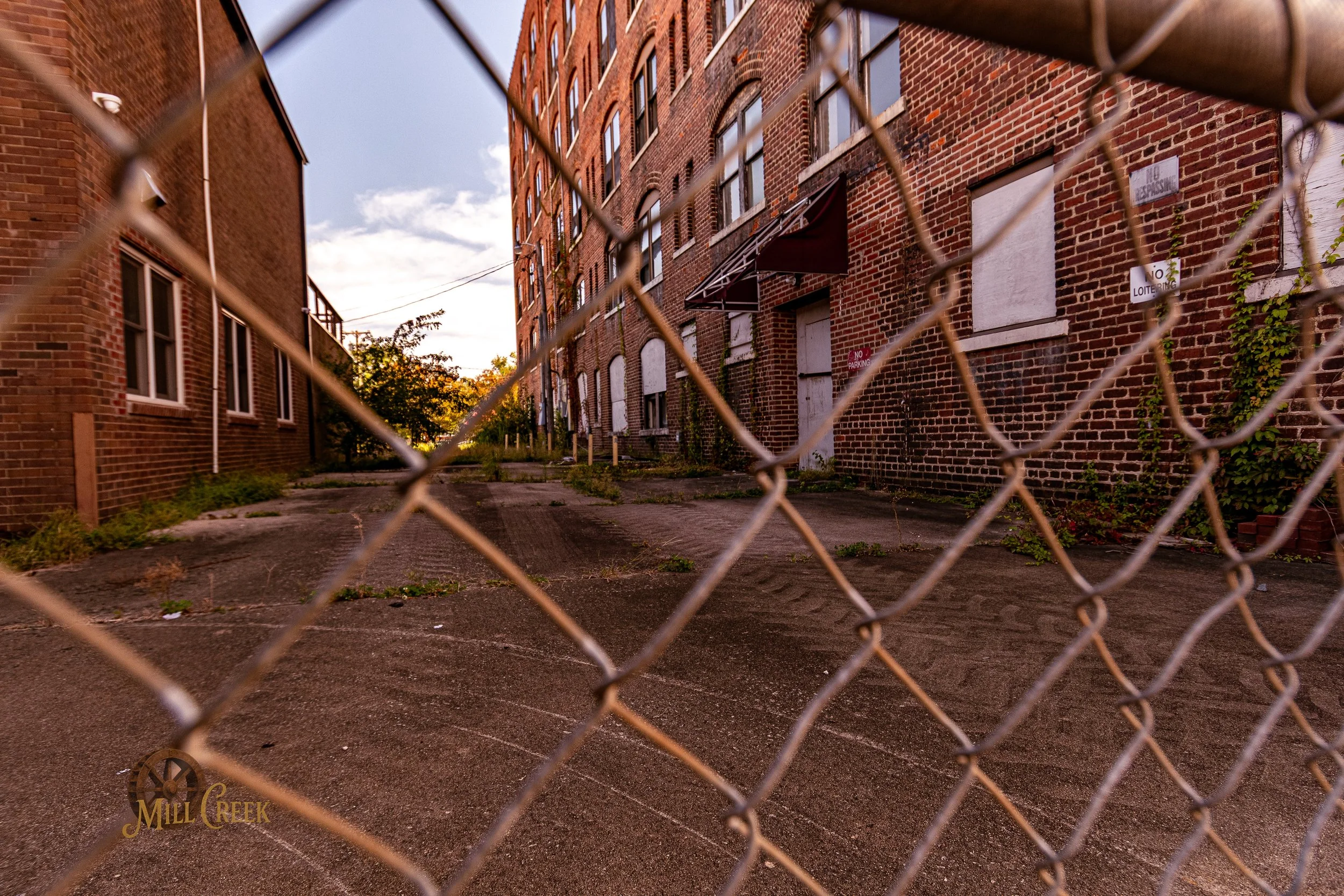 View of an alleyway through a chain-link fence, with brick buildings on both sides, some windows boarded up, and signs indicating no parking or loitering.