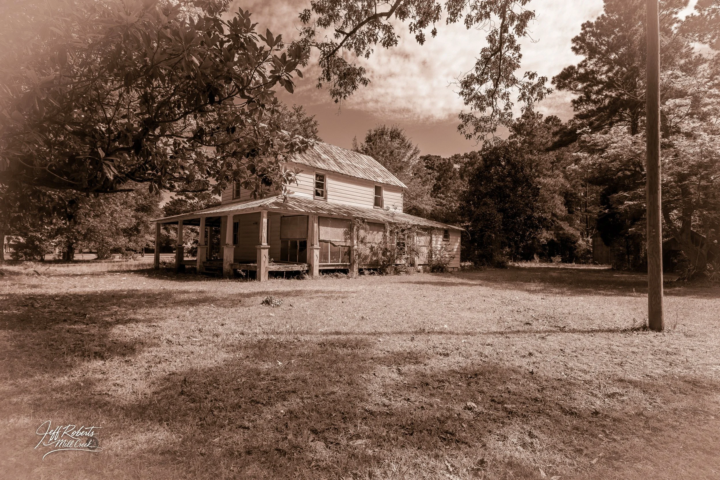 An old, abandoned two-story house with a porch, surrounded by trees and grass, overcast sky, sepia tone.