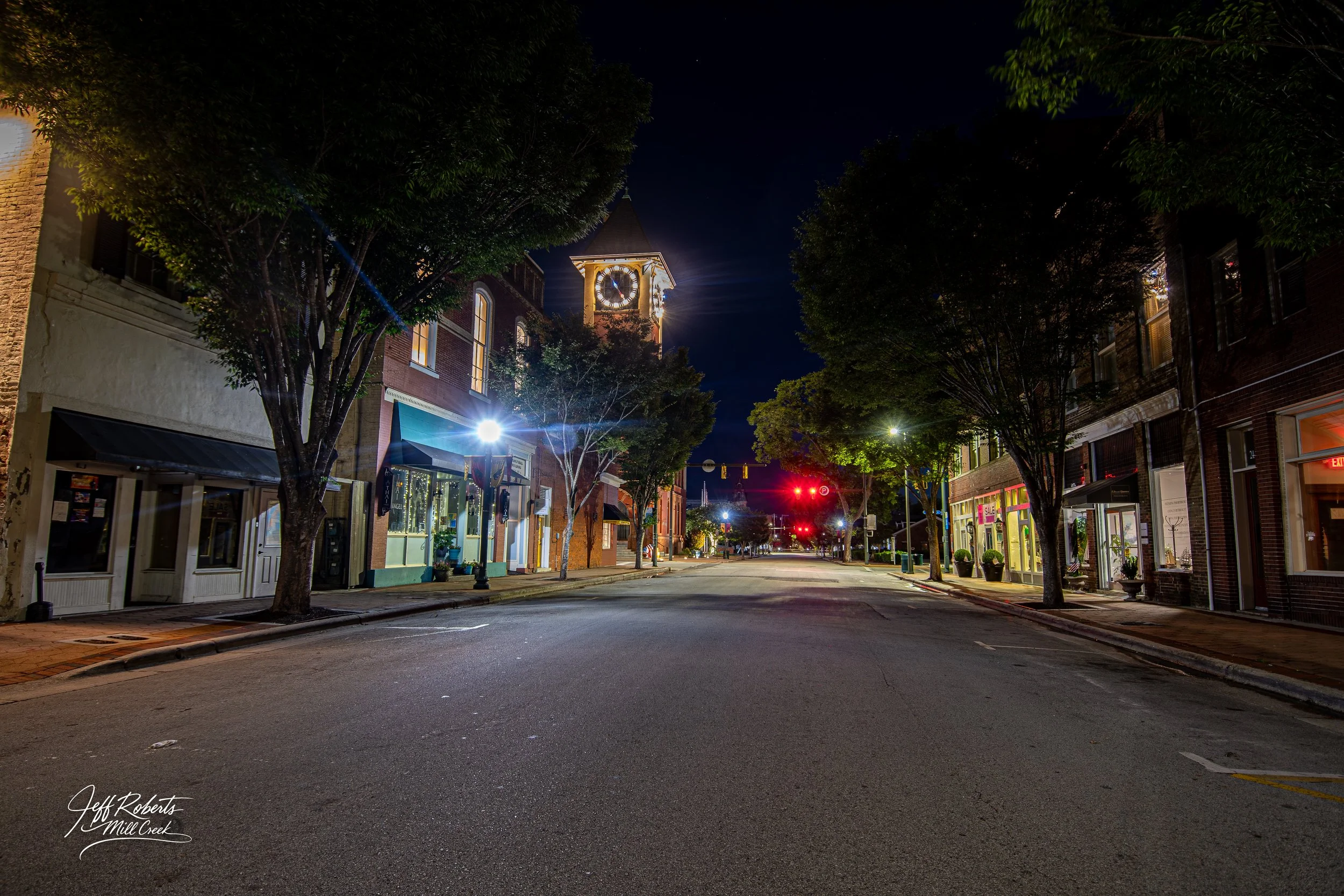 A quiet city street at night with lit buildings, trees lining the sidewalks, and a clock tower illuminated in the background.