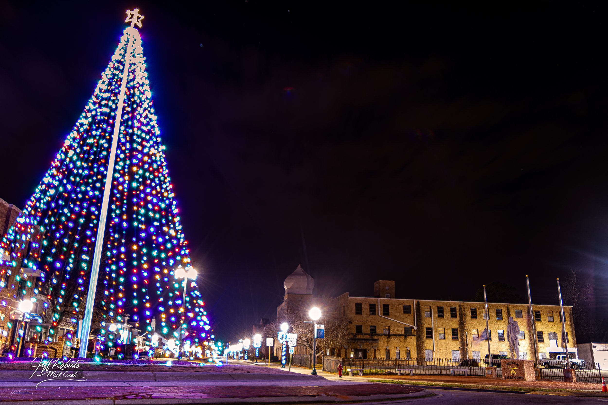 A large Christmas tree decorated with multicolored lights in a town square at night, with historic buildings, street lamps, and a statue nearby.